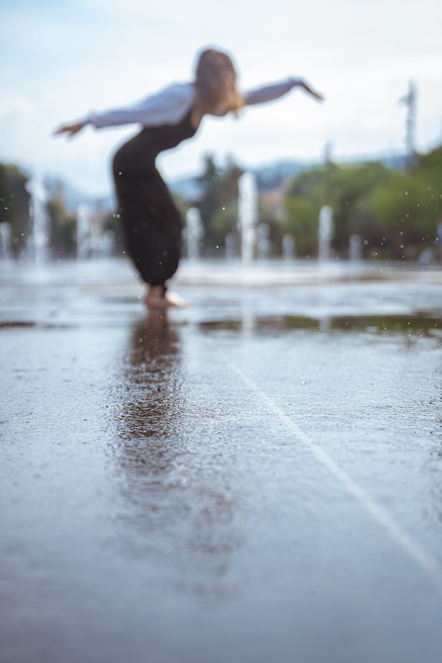 Blurred dancer balancing gracefully on wet plaza tiles surrounded by water fountain jets.