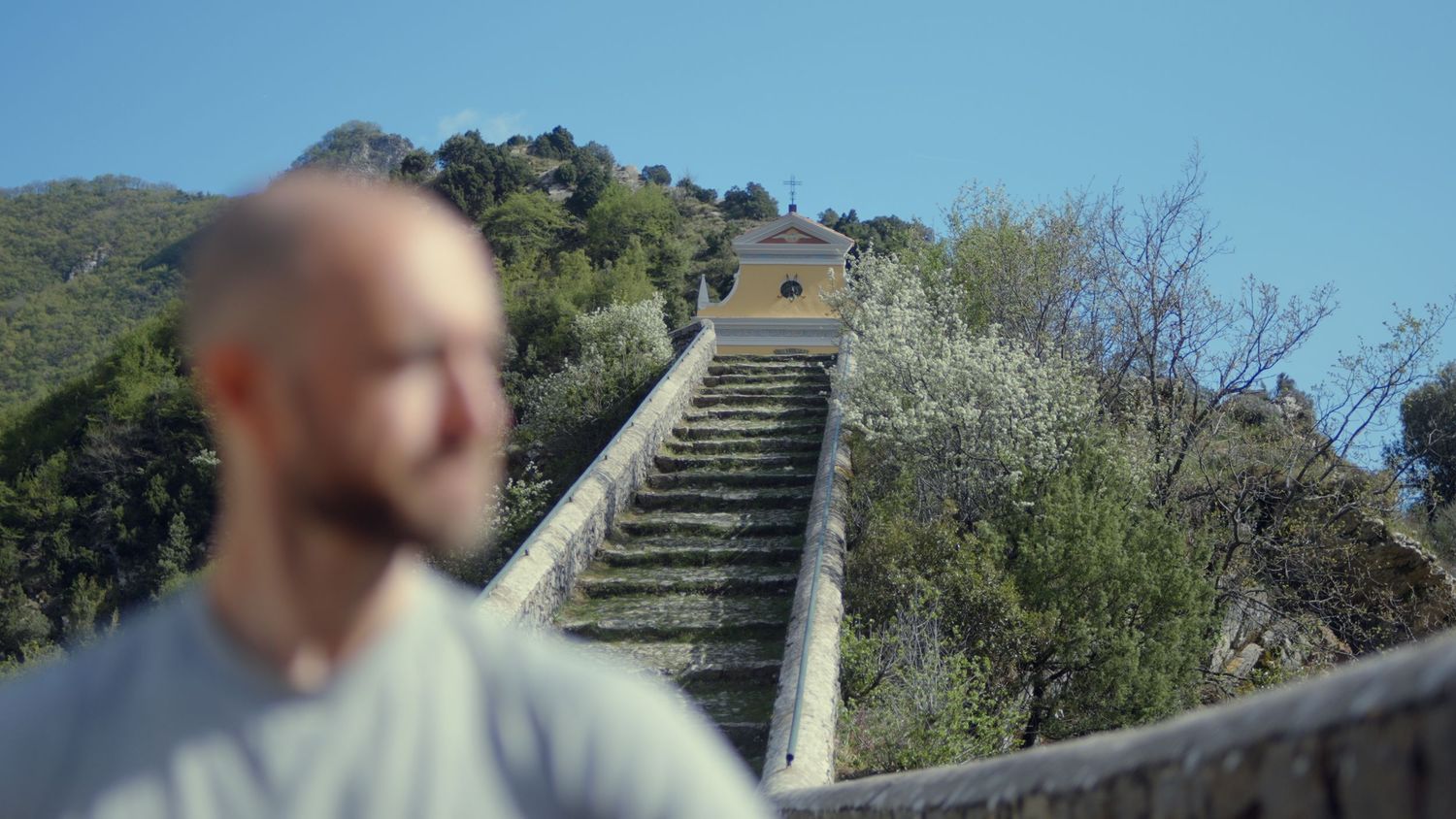 Bald man standing at base of long stone staircase on sunny hillside.
