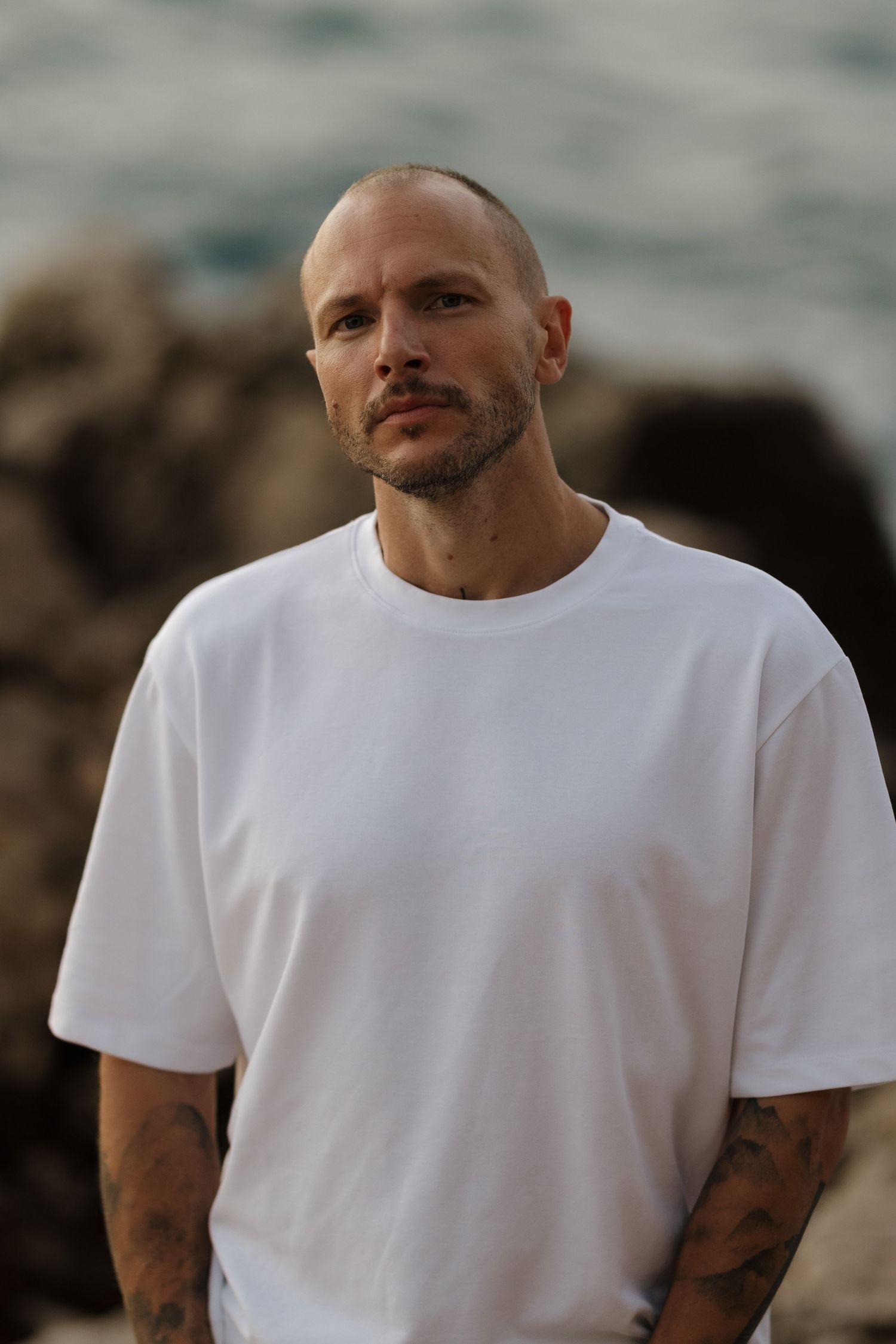 Bald tattooed man in white oversized t-shirt standing near rocky ocean shore.