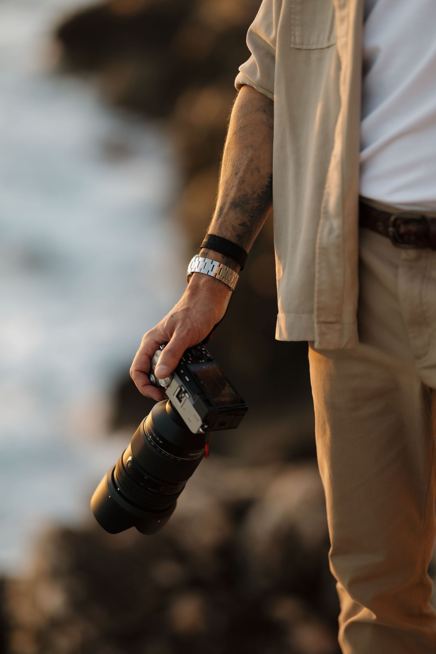 Close-up of tattooed hand holding professional camera with telephoto lens near rocky coast.