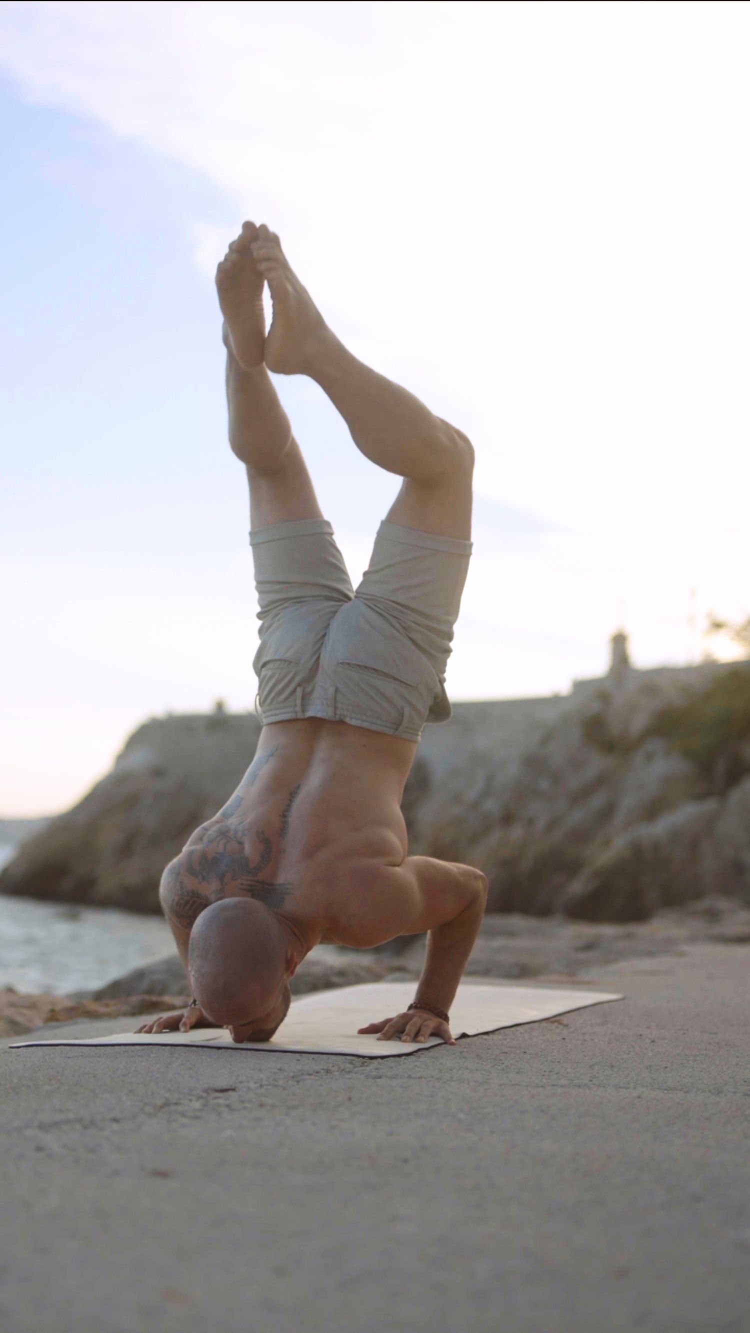 Athletic man performing a headstand yoga pose on mat near rocky beach.