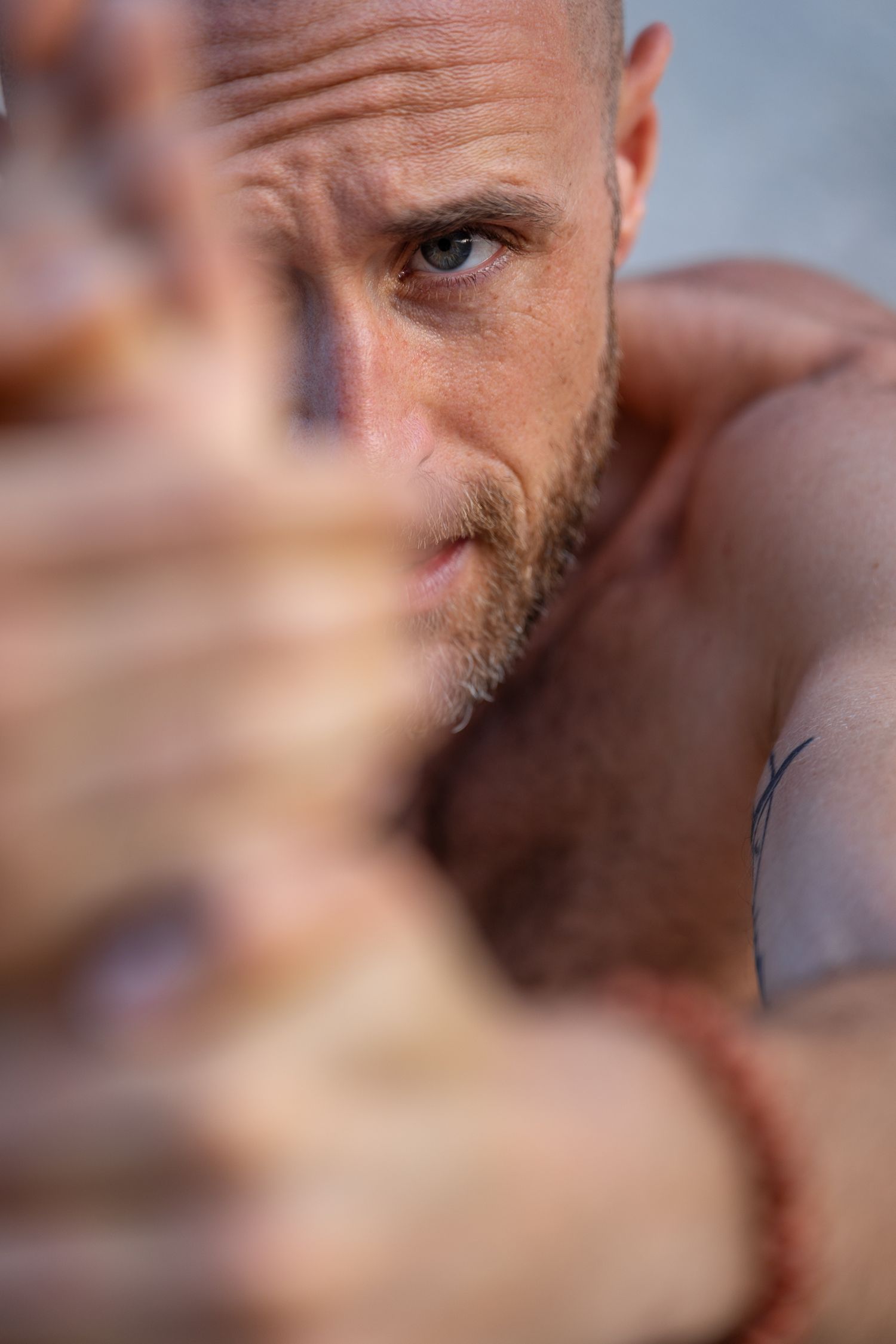 Close-up portrait of intense blue-eyed man reaching hand toward camera with tattoo visible on shoulder.