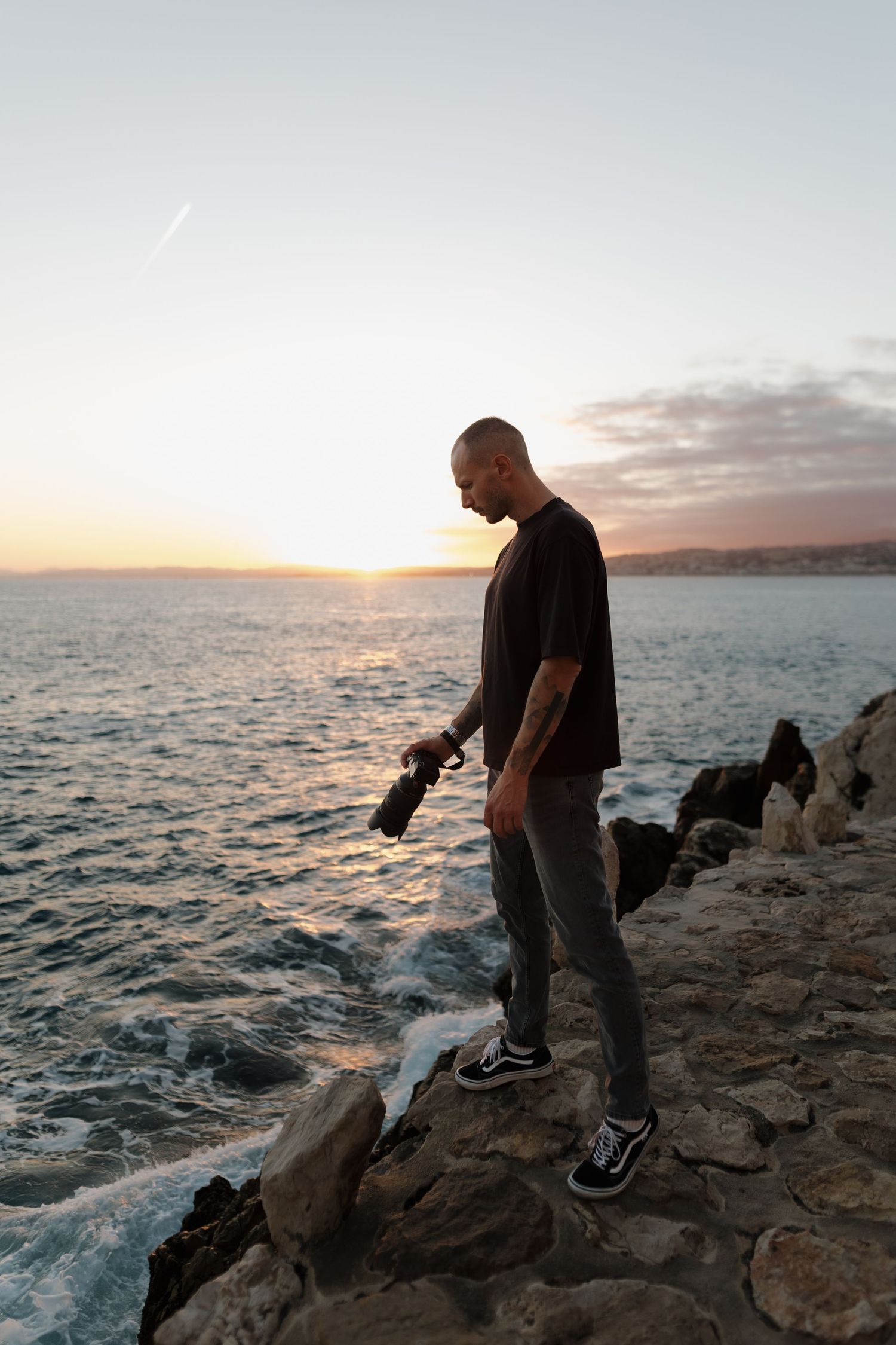 Male photographer holding camera standing on coastal rocks during golden sunset by the sea.