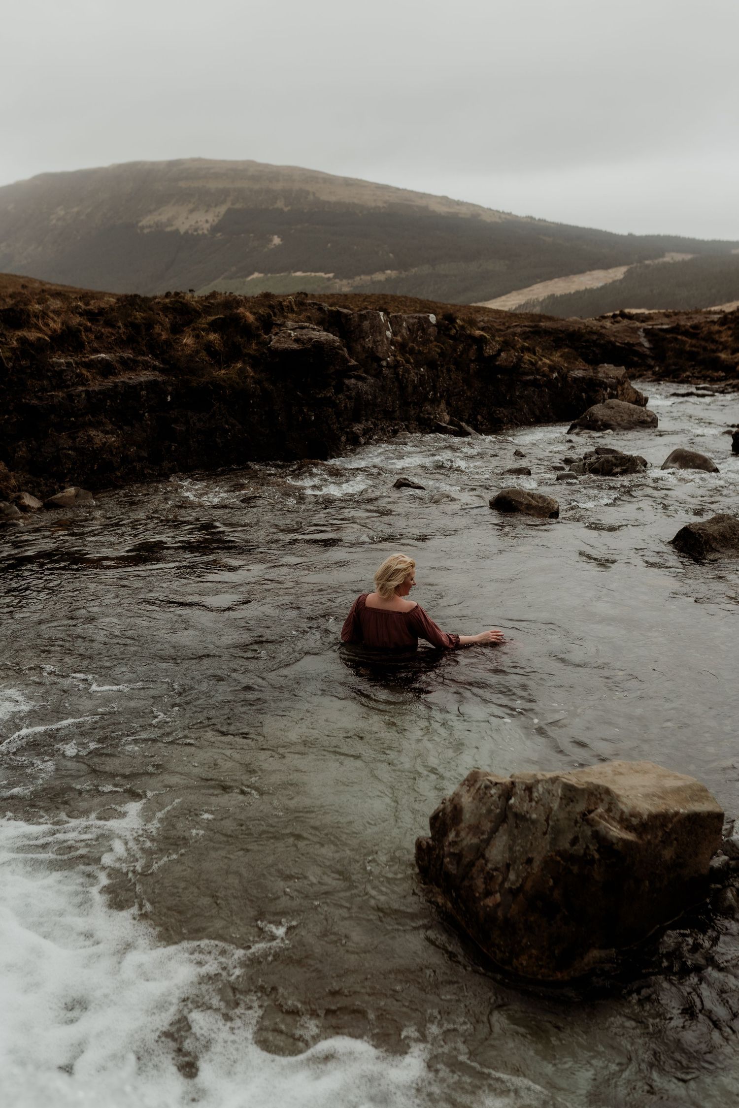 Woman sitting in a rocky natural pool with misty mountain scenery on an overcast day.