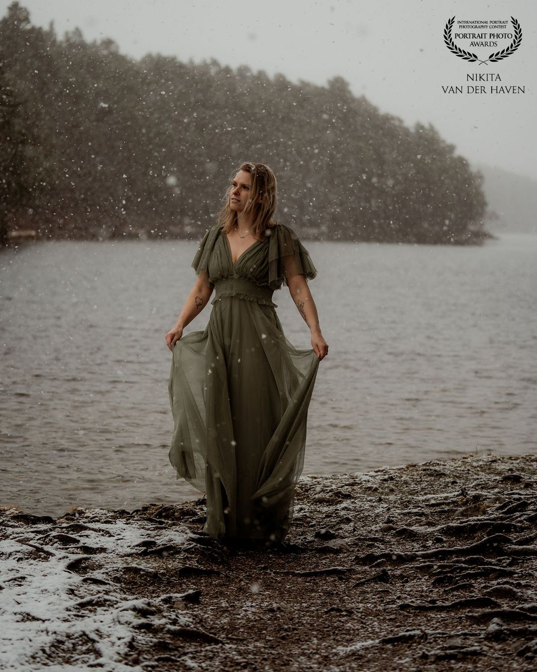 Woman in green dress standing in shallow water during snowfall with misty forest background.