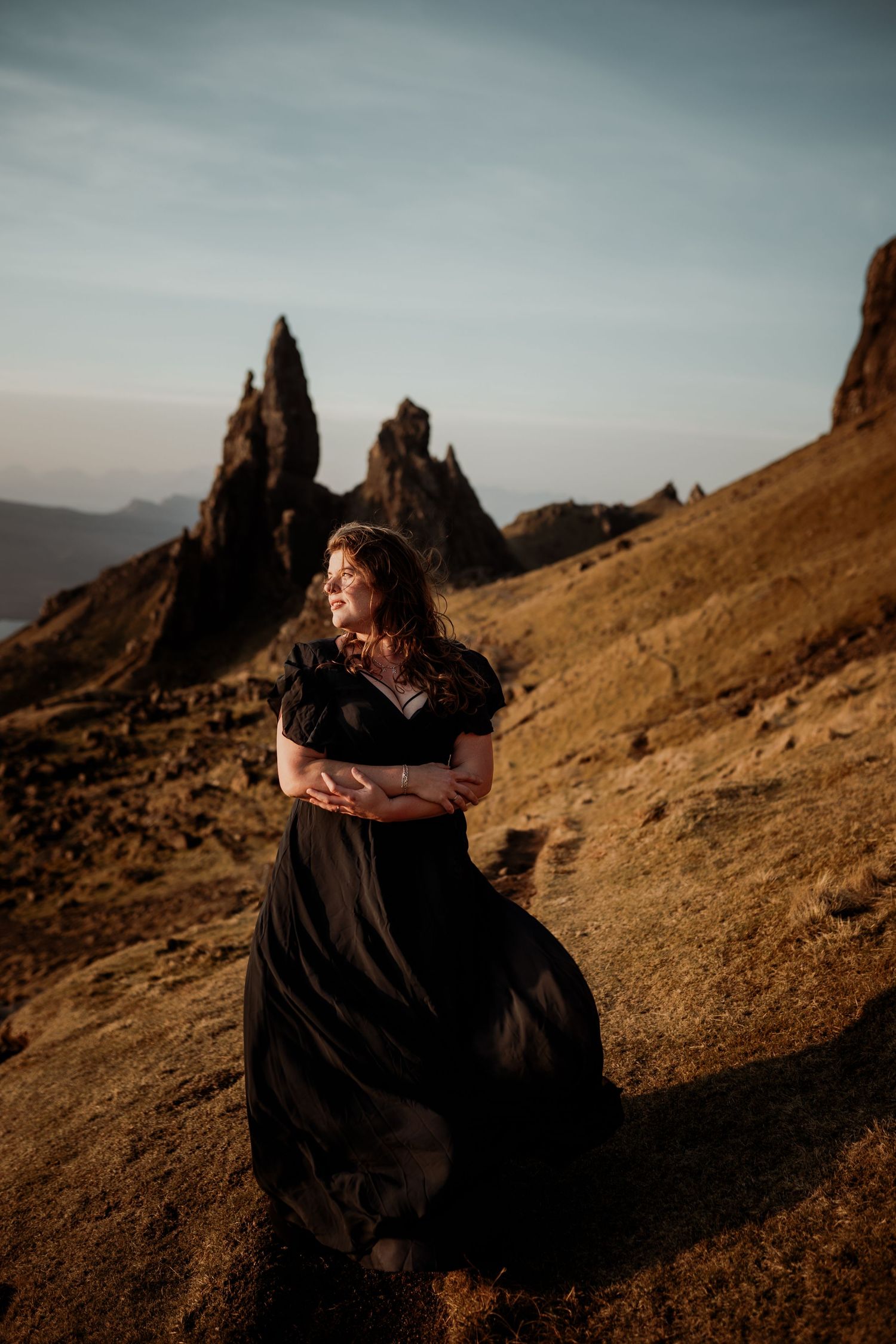 A flowing black dress catches the wind against dramatic rocky spires of the Old Man of Storr in Scotland at sunset.