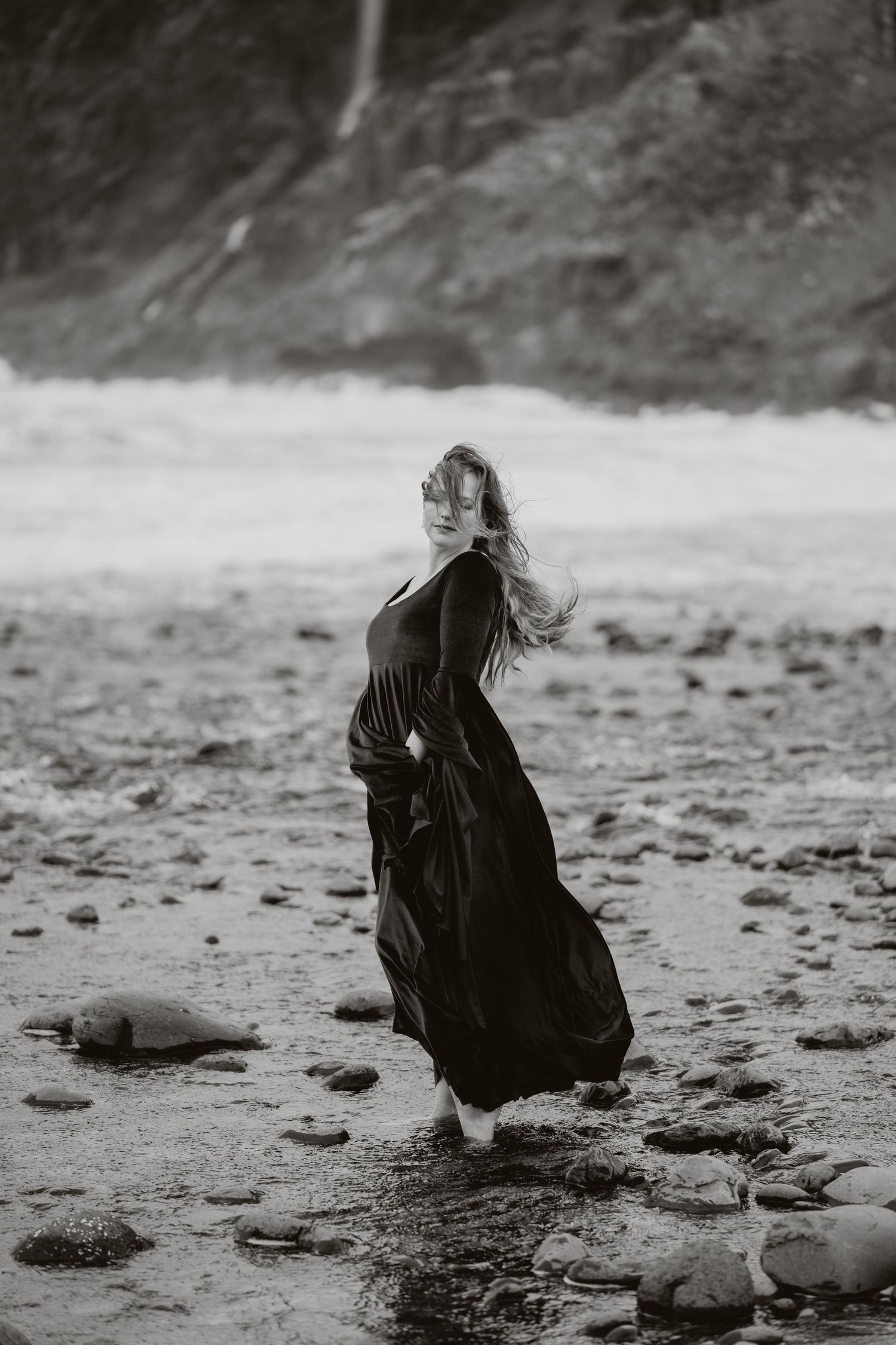 A woman in a flowing black dress stands in a rocky stream with wind-blown hair in a dramatic black and white photo.