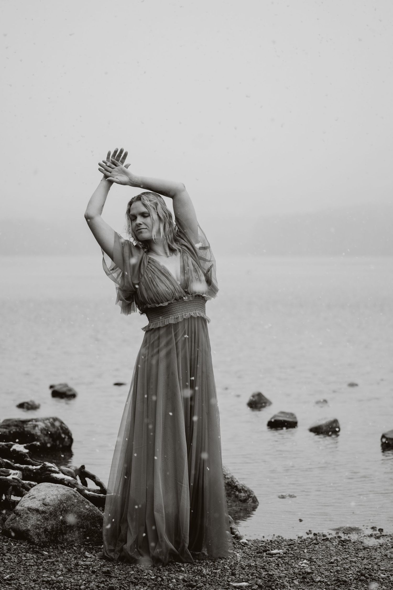 Woman in flowing gown poses gracefully on rocky shoreline in moody black and white photography.