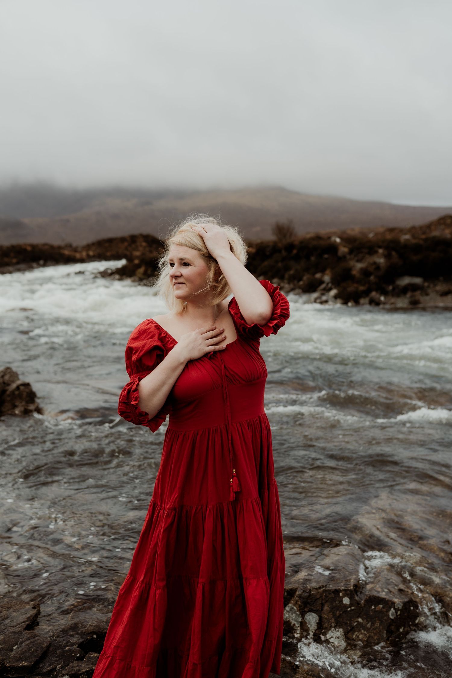 Woman in red dress standing by a rushing river with misty mountains in the background.