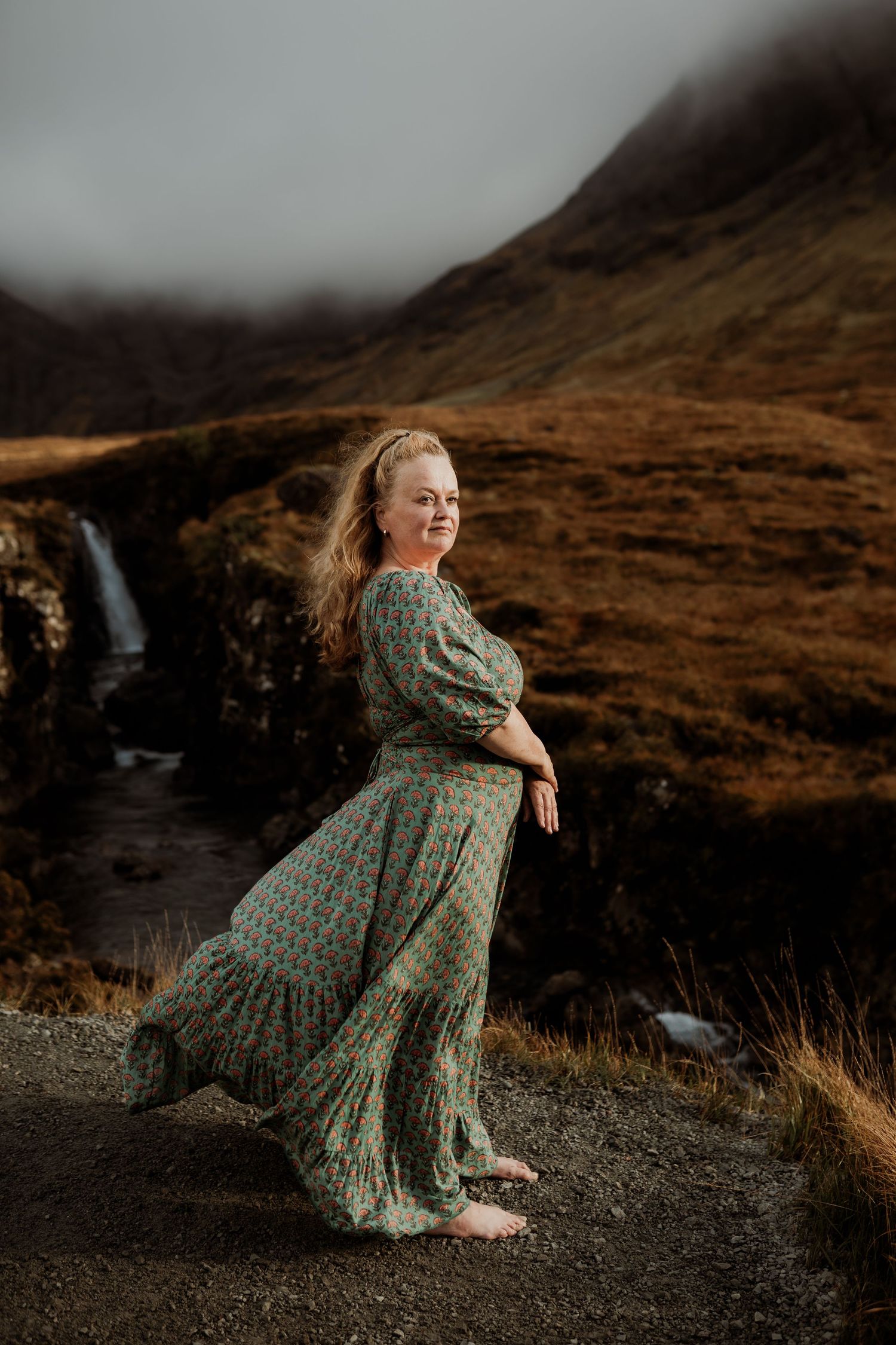 Person in a flowing green dress twirls against a scenic backdrop of Scottish highlands with a cascading waterfall.