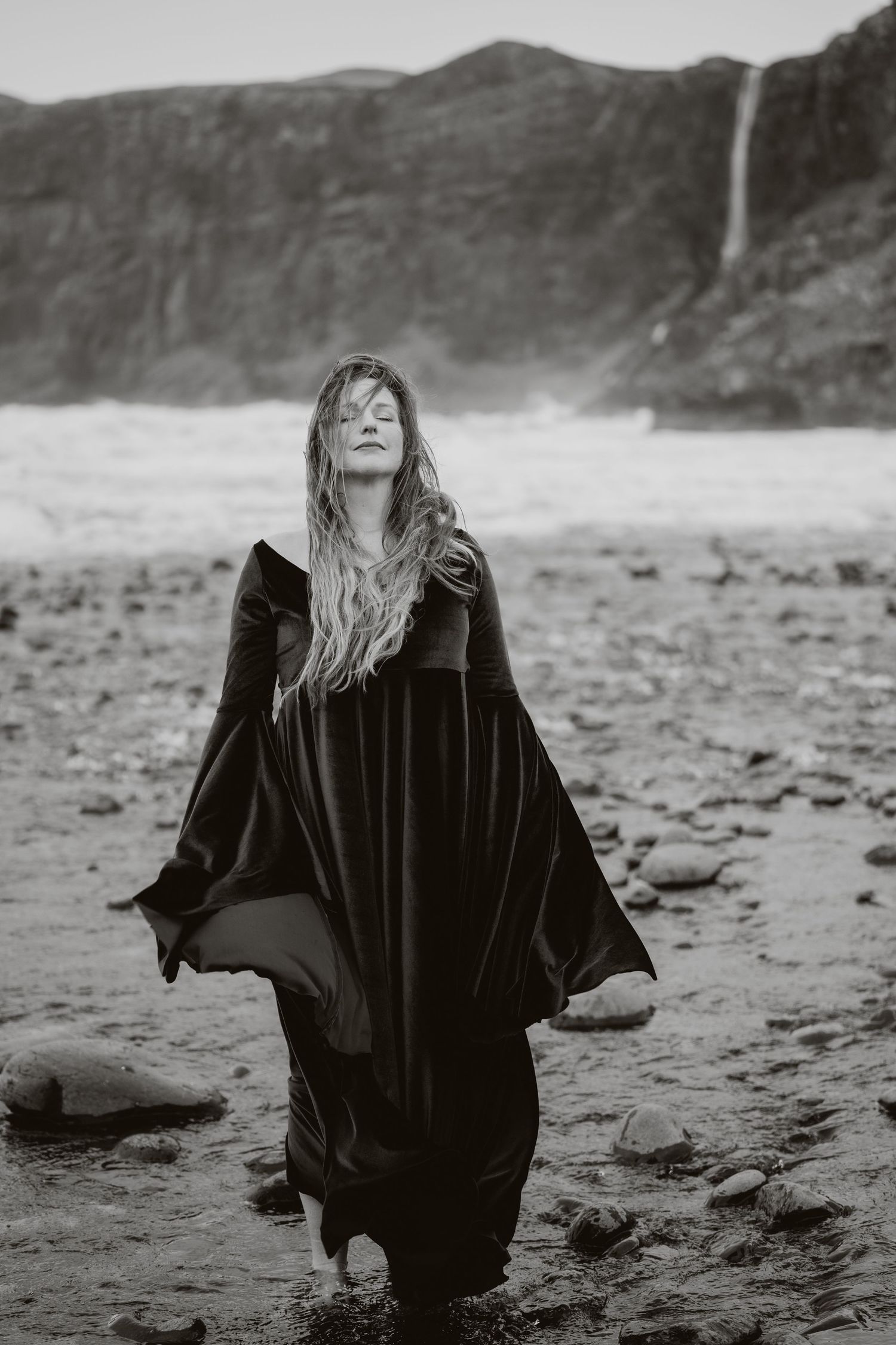 Woman in black medieval gown standing in rocky shoreline waters with misty cliffs and waterfall backdrop.