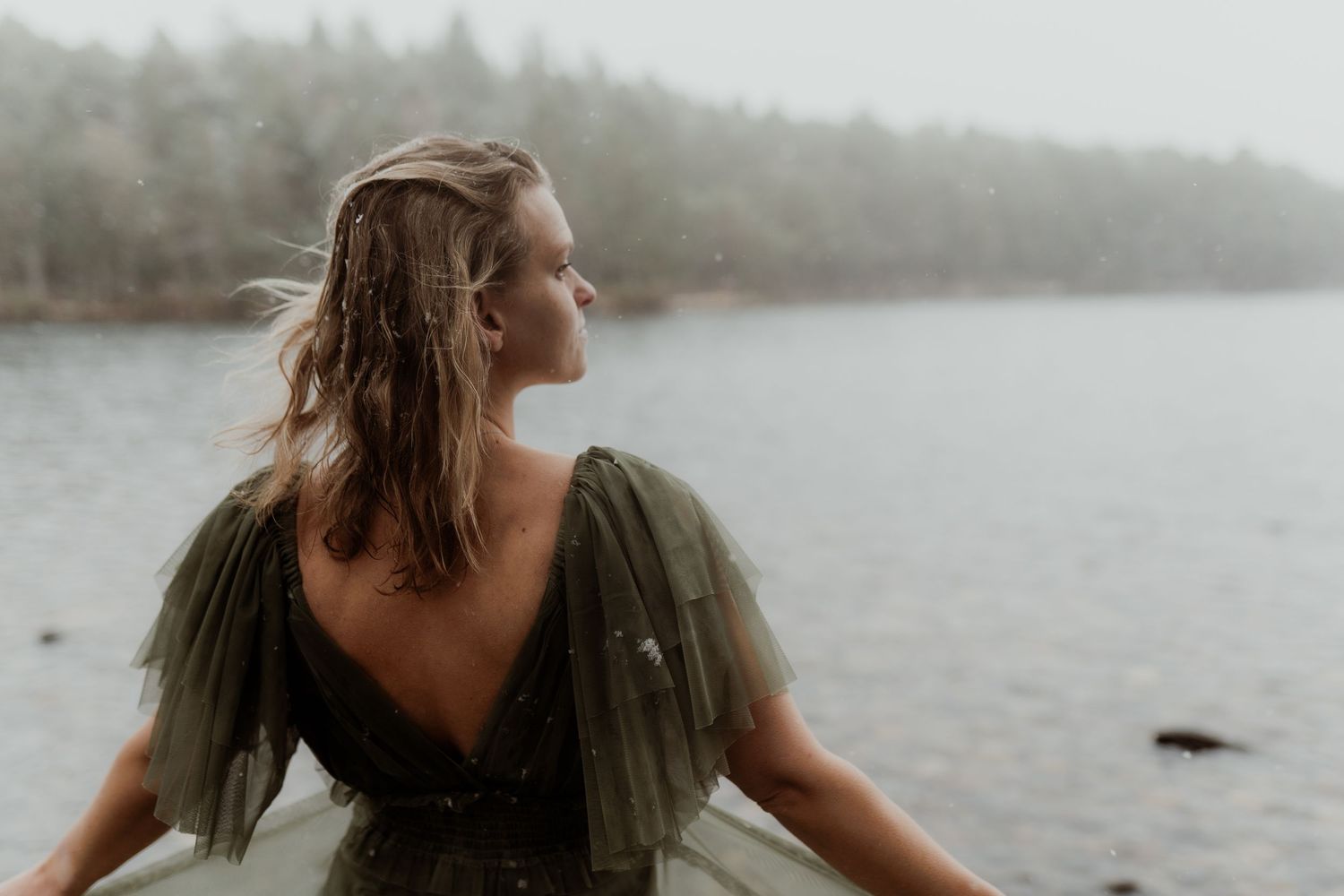 Woman in olive tulle dress stands by misty lake with wind-blown hair.