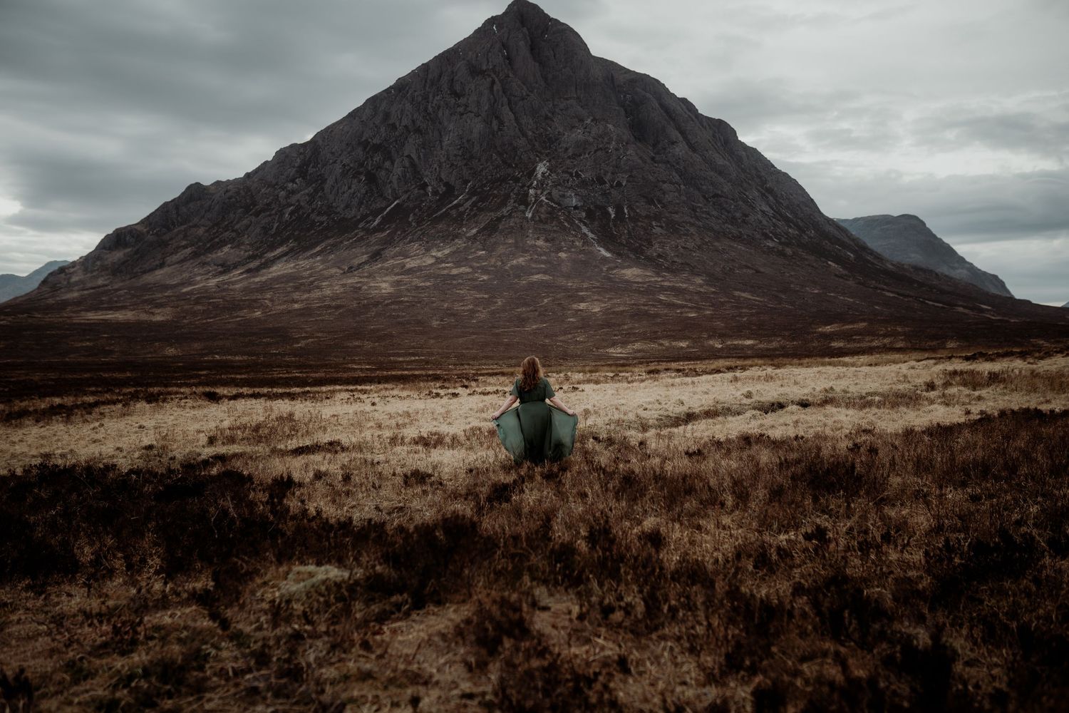A figure in a green coat stands in the rugged Scottish Highlands beneath the dramatic Buachaille Etive Mor mountain.