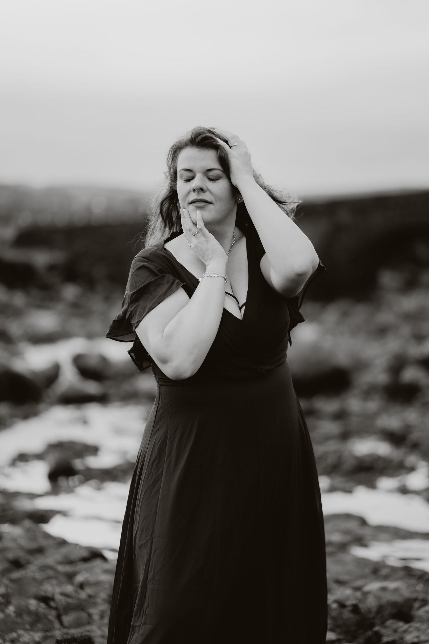 Artistic black and white portrait of person in dark dress with hands near face against moody outdoor landscape.