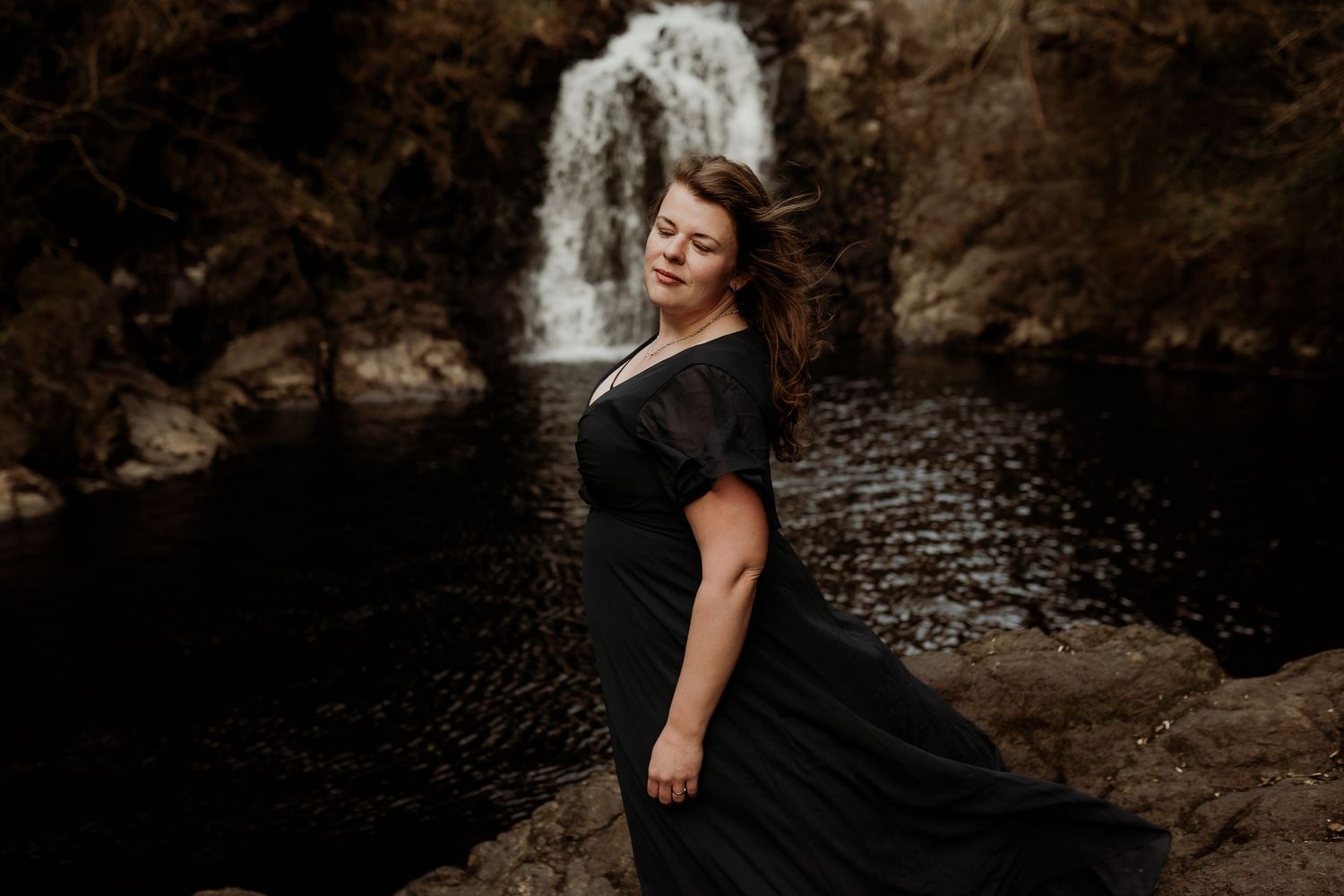 Female in black dress poses dramatically by scenic waterfall in moody nature setting with rocky landscape.