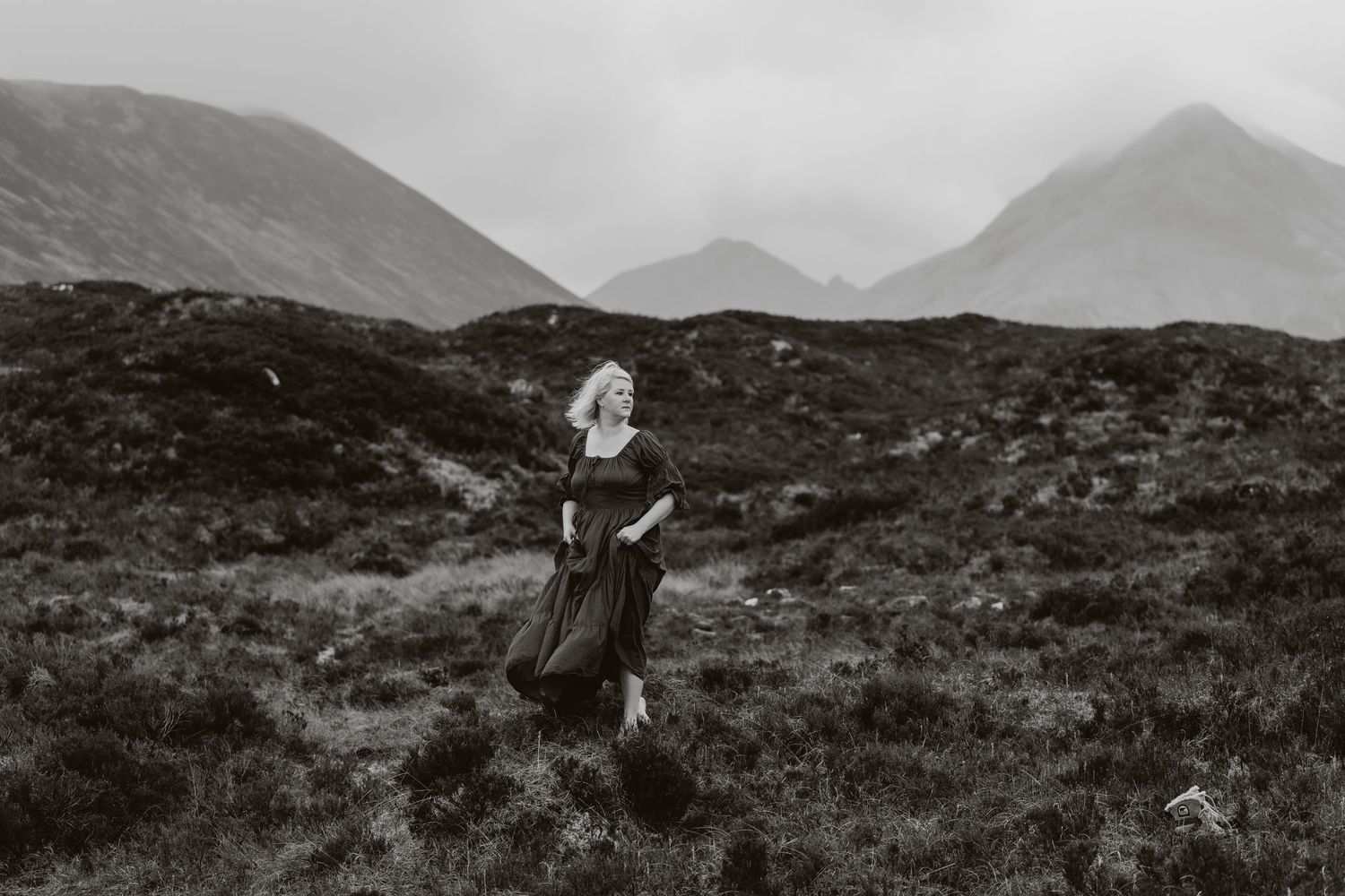 Woman in black dress walking through misty highland moorland with dramatic mountain backdrop in black and white.