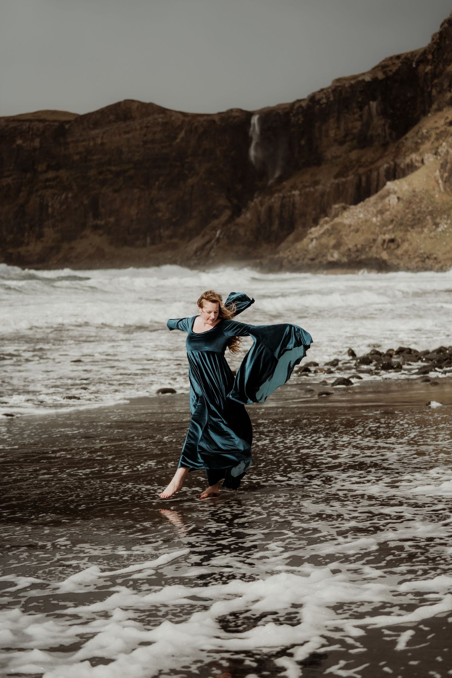 Woman in teal dress dancing on rocky beach with ocean waves and dramatic cliffs in background.