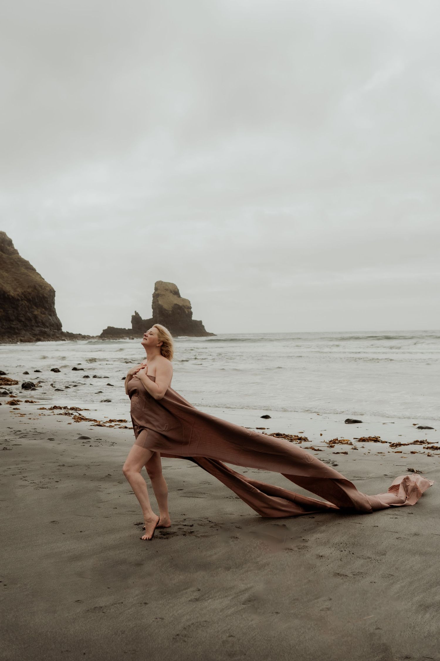 Woman wrapped in flowing brown fabric poses on a misty beach with rocky sea stacks in the background.