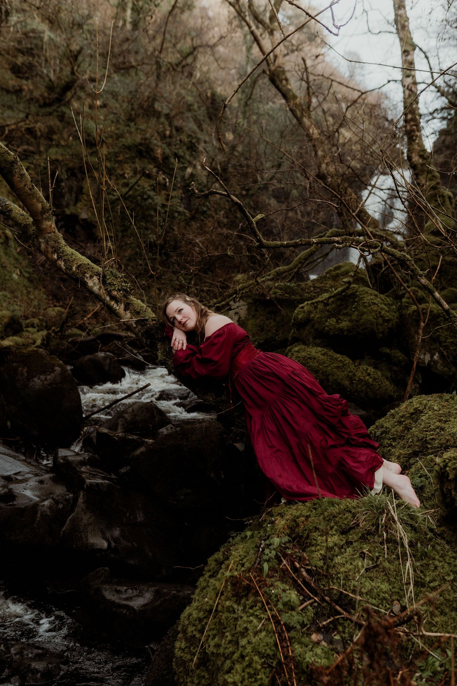 Woman in red gown reclining on mossy rocks beside a forest stream.