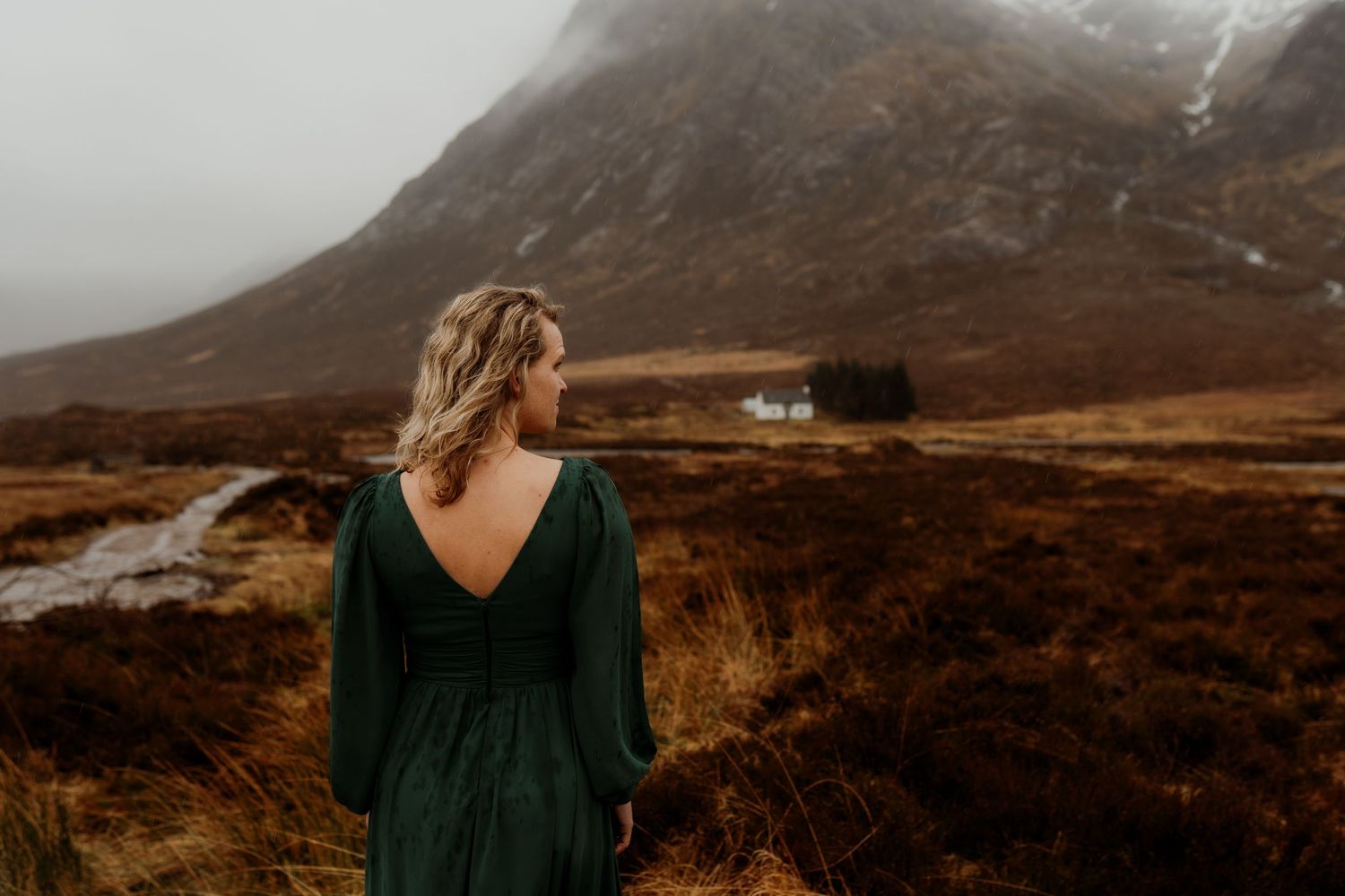 Woman in green dress gazes at misty Scottish highlands landscape with white cottage.