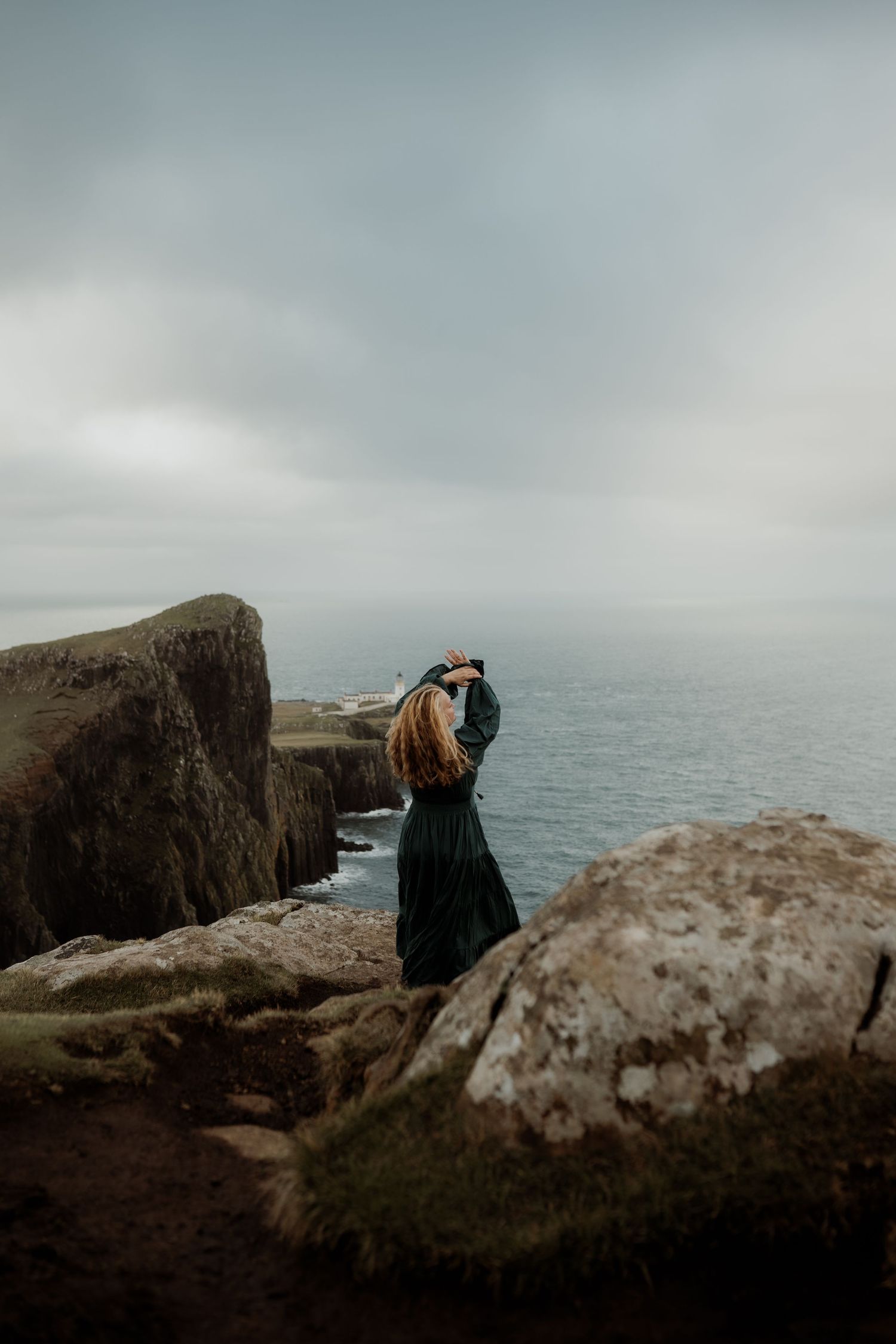 Two figures stand at the edge of dramatic coastal cliffs overlooking a misty ocean on a moody, overcast day.