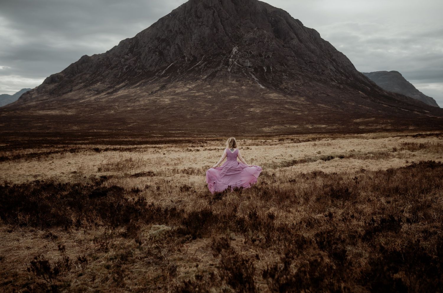 Figure in flowing purple dress stands alone on misty Scottish moorland beneath dramatic mountain peak in moody lighting.