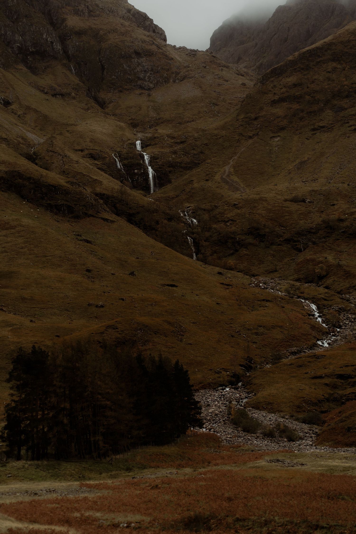 A dramatic waterfall cascades down rugged brown mountainside in Scottish Highlands with dark pine trees at the base.