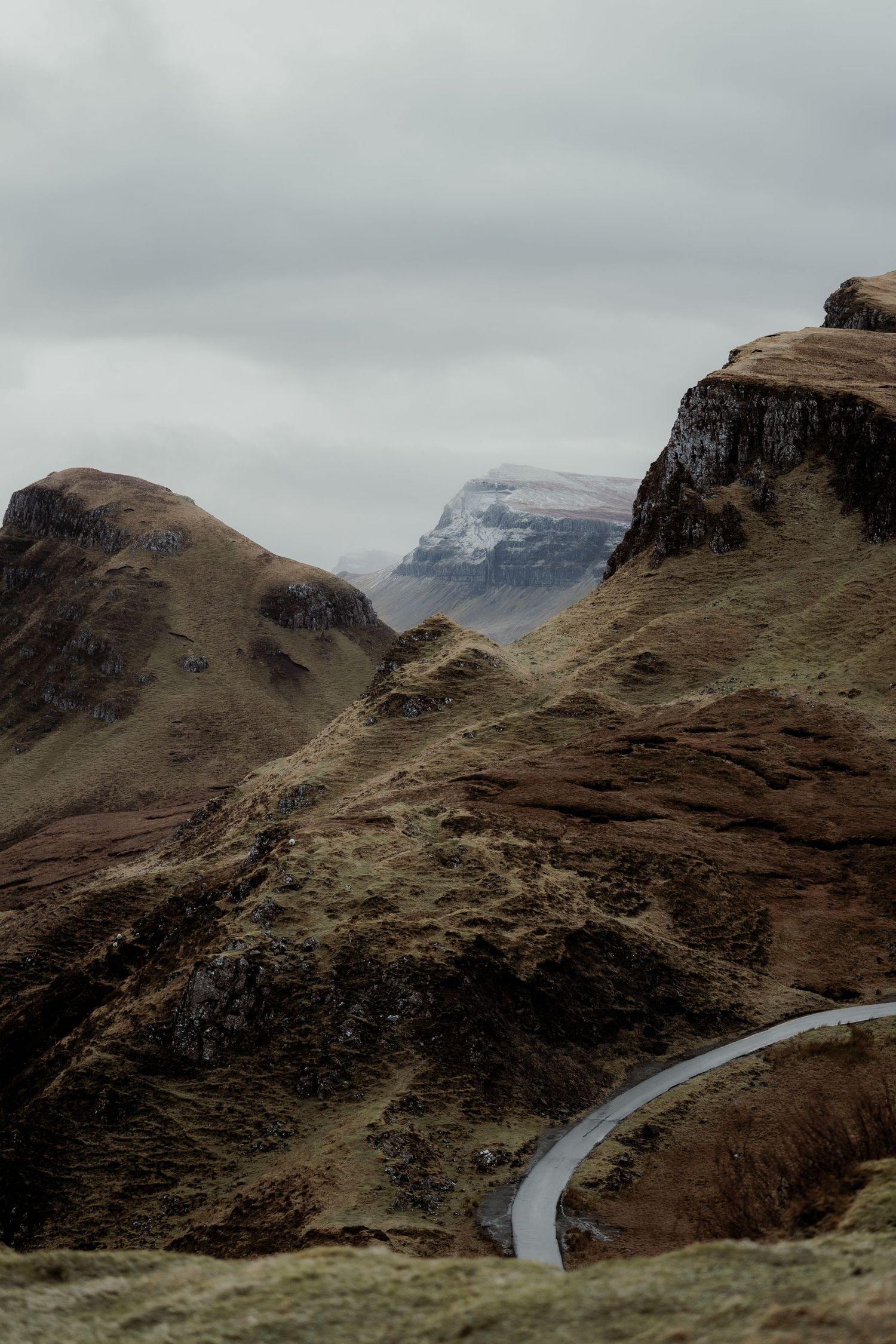 A winding road curves through dramatic rocky highland mountains under a cloudy grey sky.