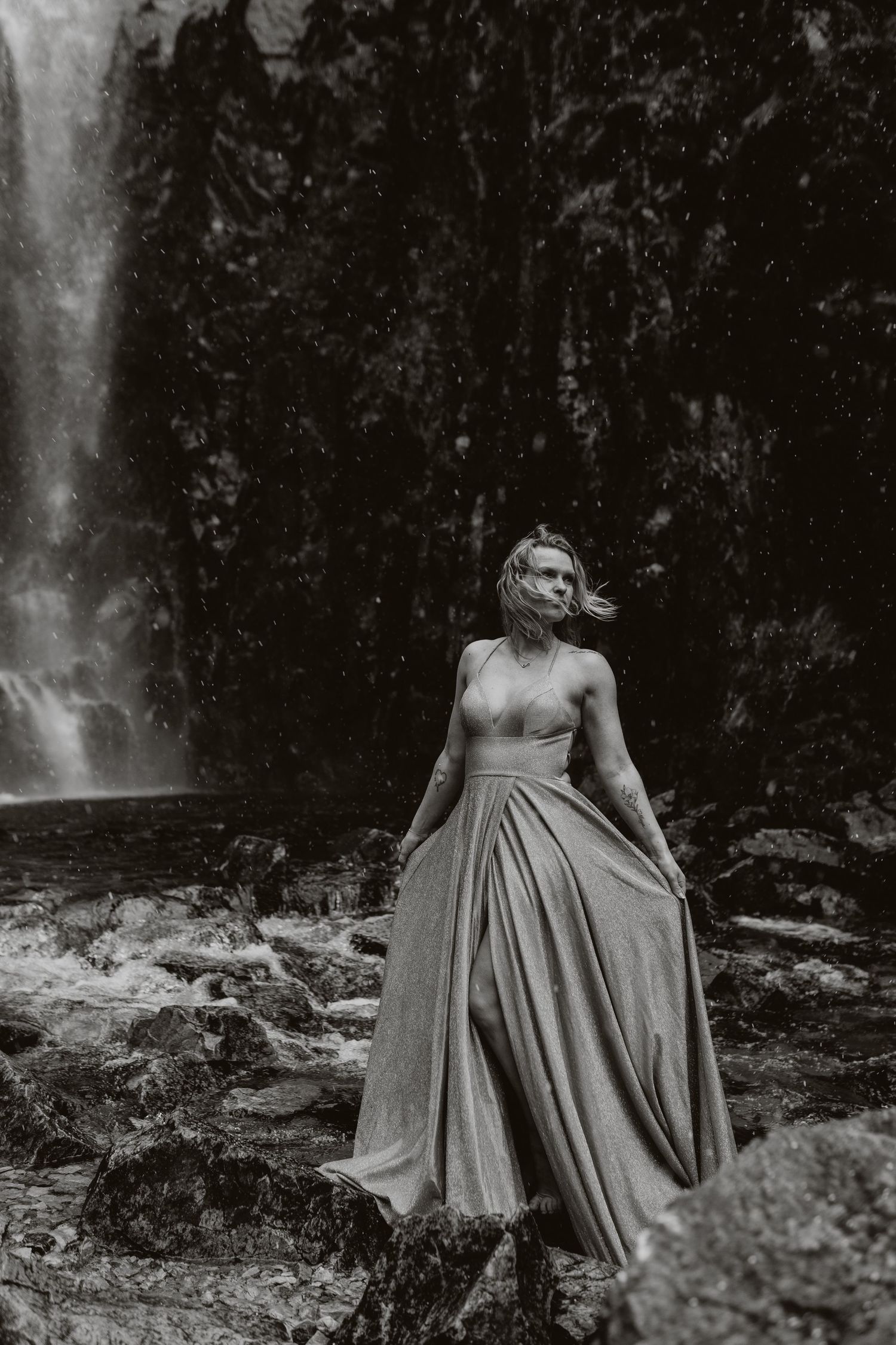 Black and white photo of a woman in an elegant gown standing before a waterfall on rocky terrain.
