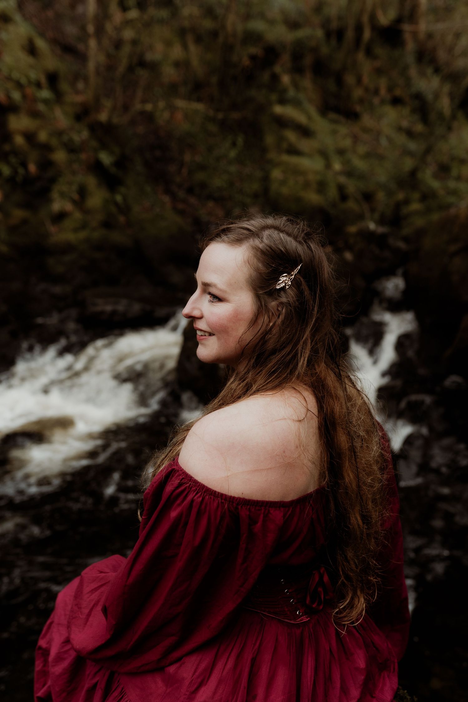 A woman in a red off-shoulder dress sits smiling beside a rushing waterfall in lush greenery.