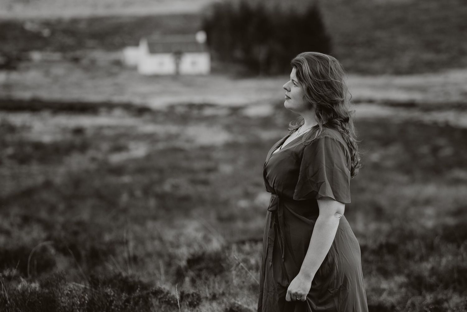 A moody black and white portrait of someone in a dress standing in a highland landscape with a white cottage in the distance.