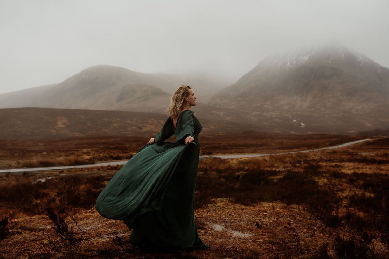 Woman in flowing green gown poses dramatically against misty Scottish Highland mountains.