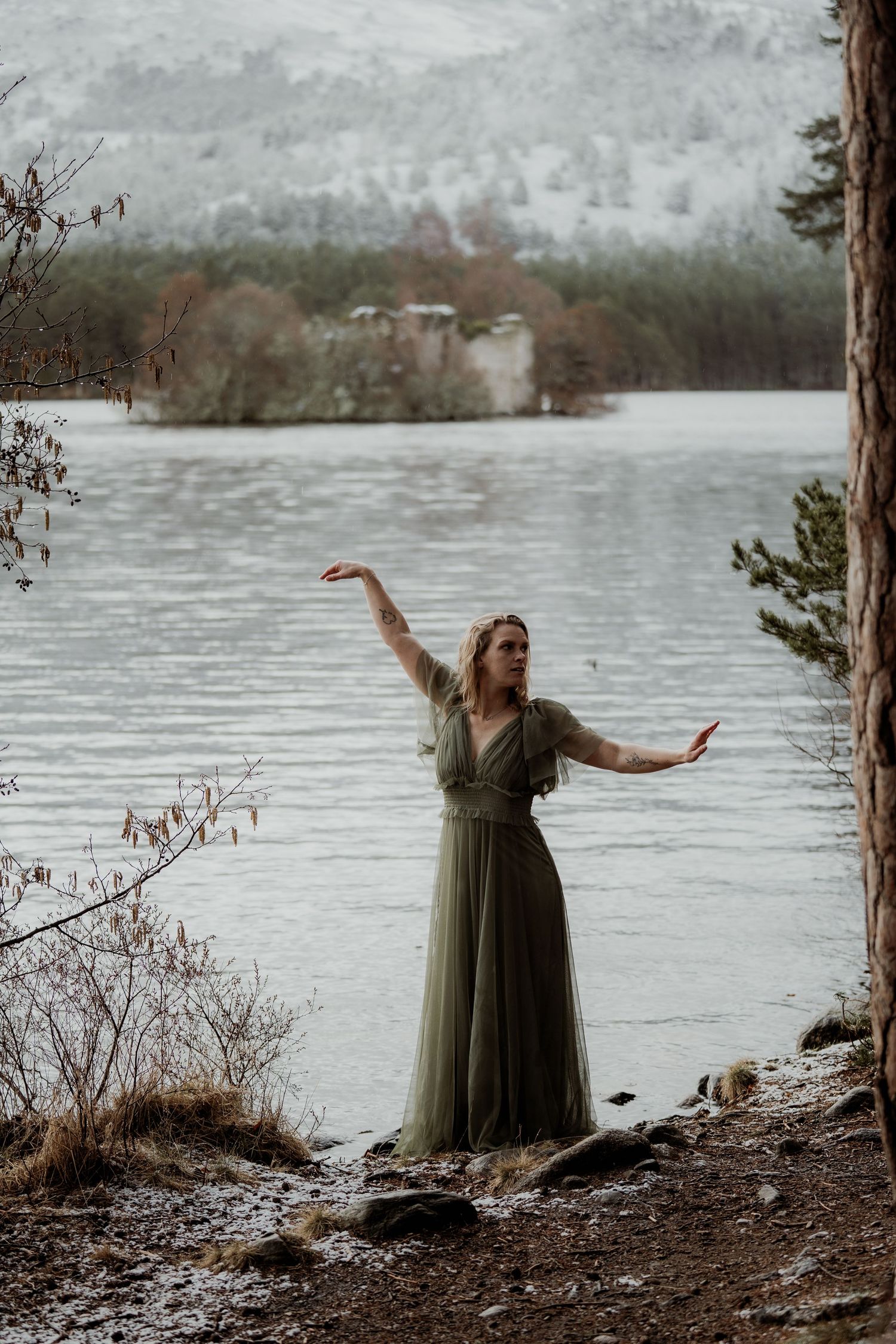 Woman in elegant olive green gown posing gracefully by a misty winter loch with snowy hills.