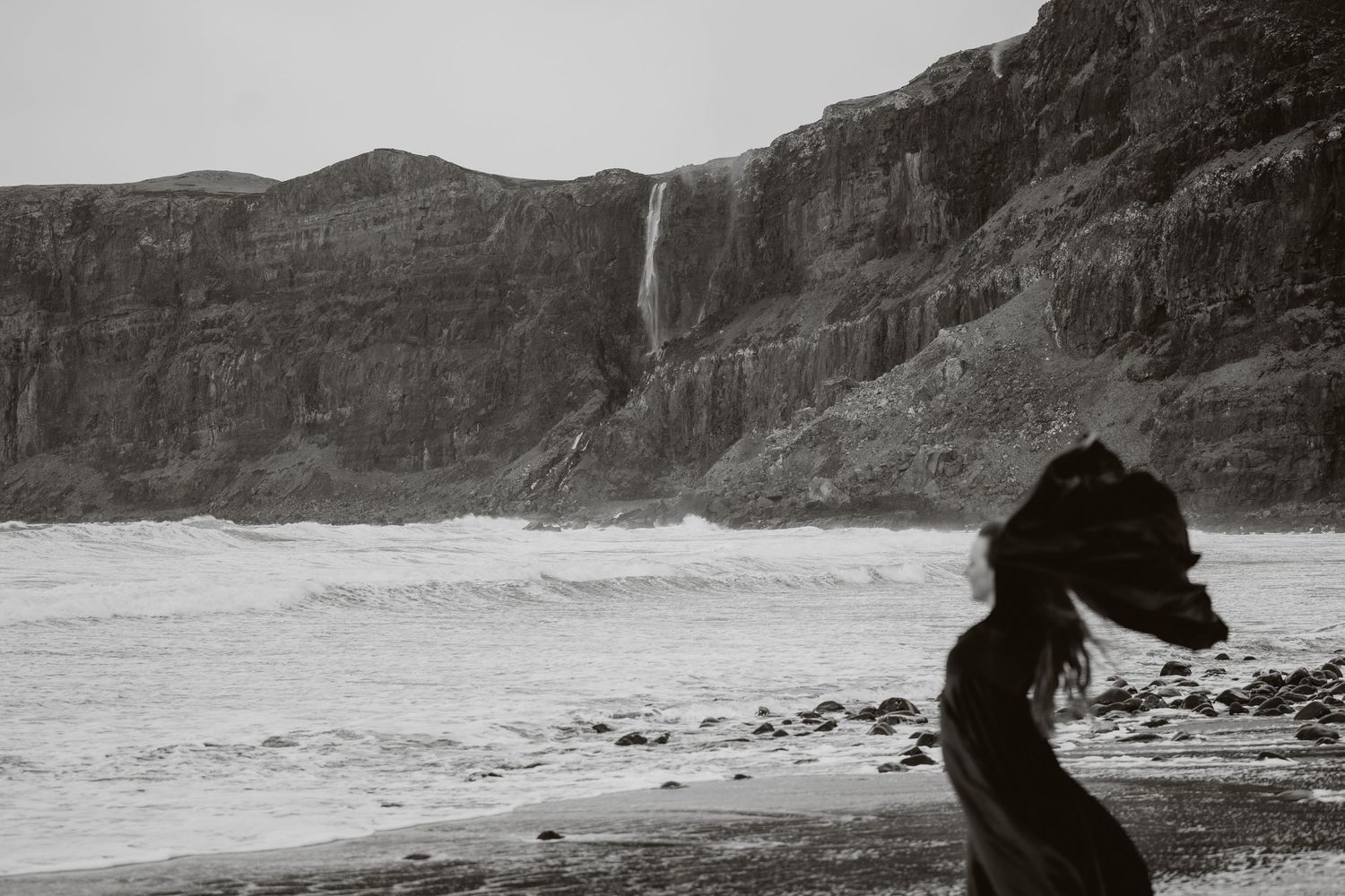 Black and white photo of a woman on a rocky beach with dramatic cliffs and a waterfall in the background.