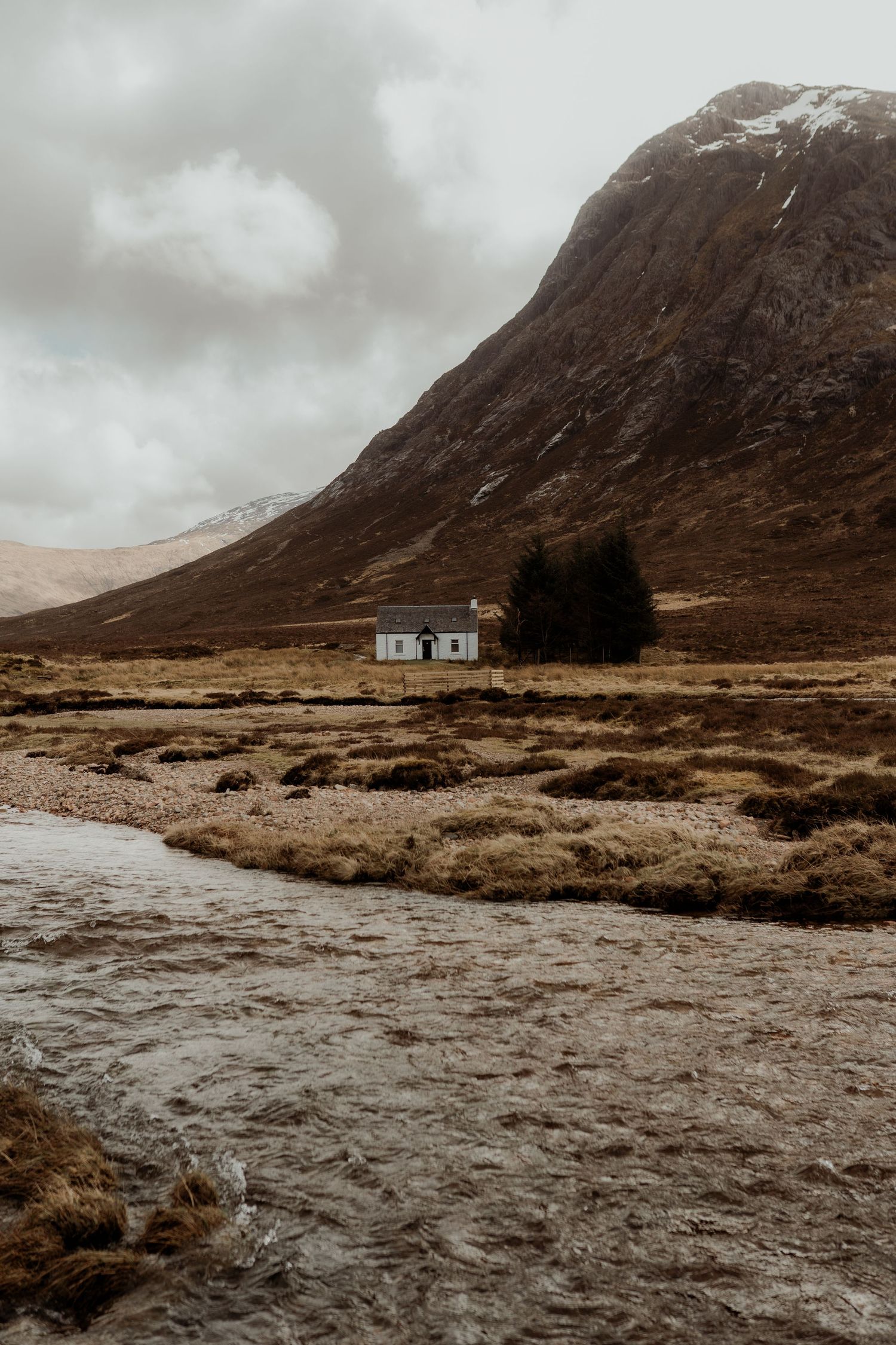 Lone white cottage beside trees in a vast Scottish Highland valley with a rocky river and snow-capped mountain under cloudy skies.