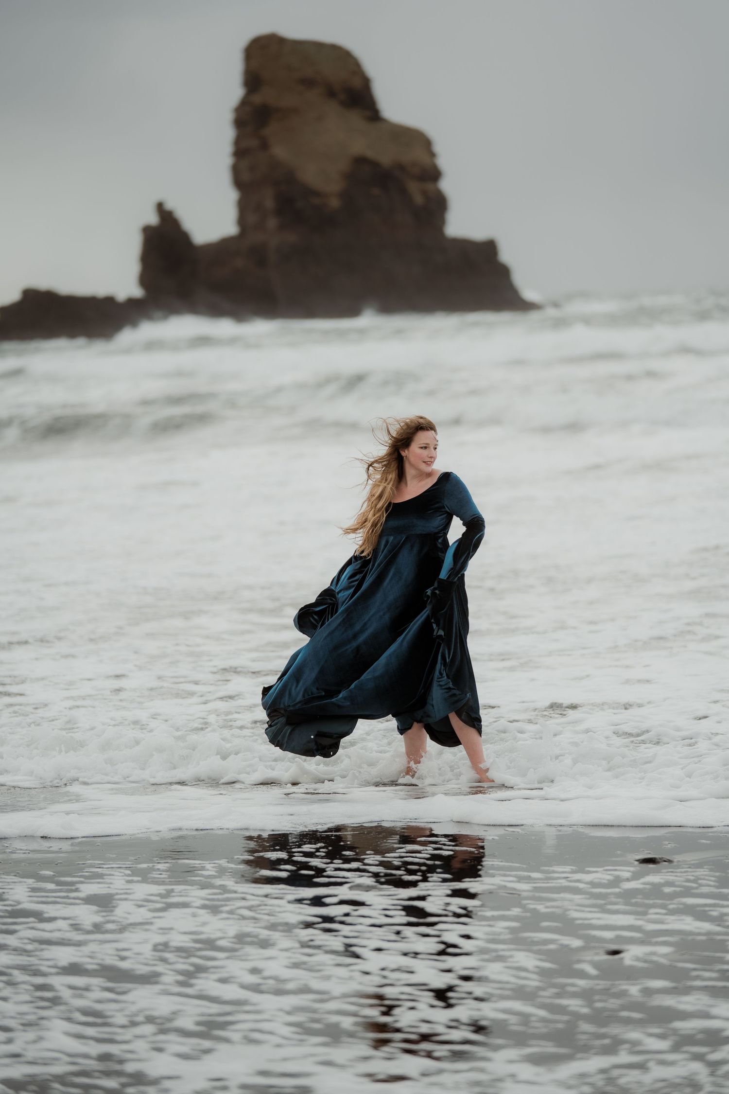 Woman in dark blue velvet gown walking through ocean waves on a stormy beach with sea stacks.