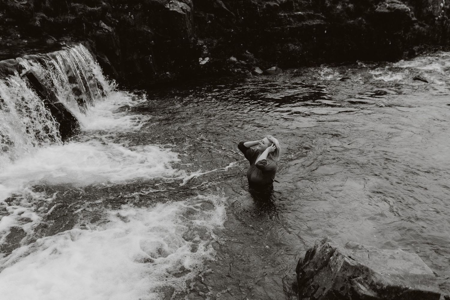 Person wading in rocky river near small waterfall, black and white nature photography.
