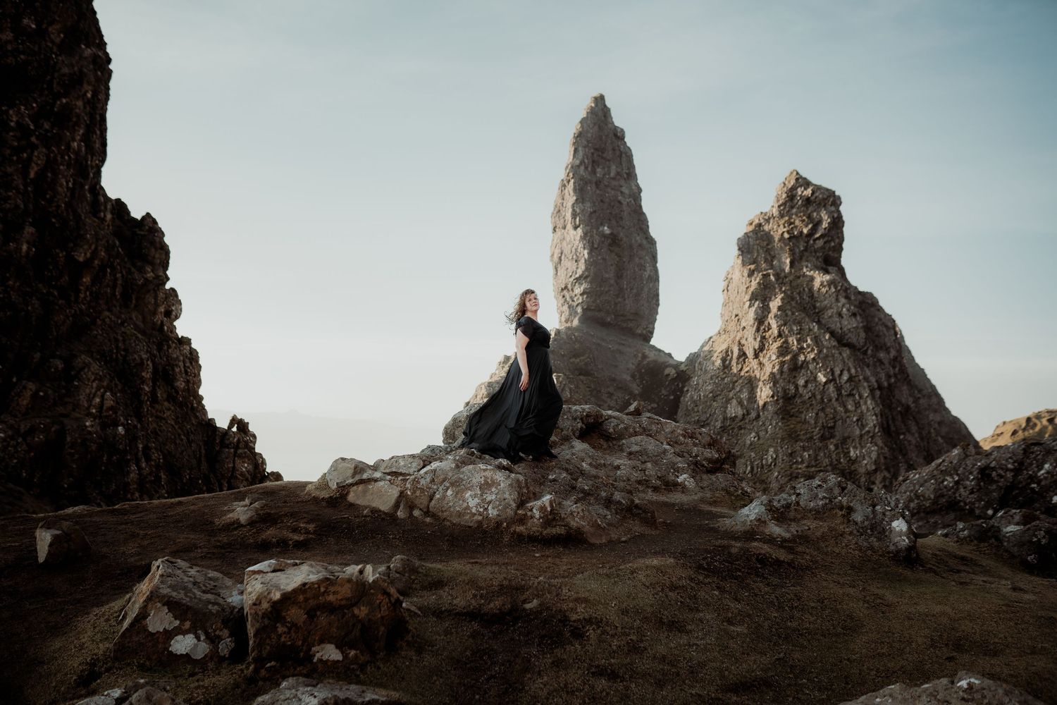 A silhouette sits on rocky cliffs near the towering Old Man of Storr formation on Isle of Skye in Scotland at sunset.