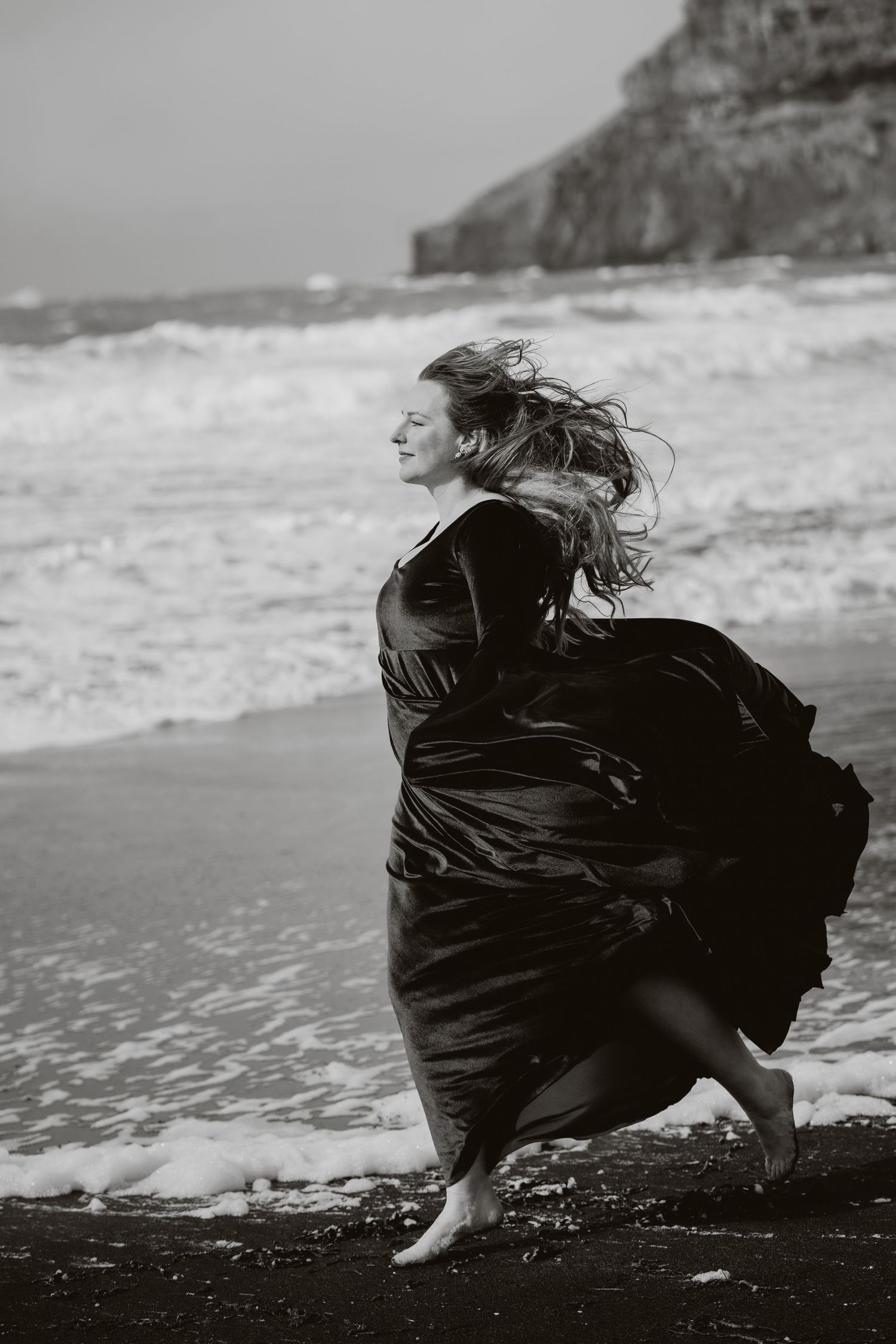 Black and white photo of a woman in a flowing dark gown running barefoot along a rocky beach with waves crashing behind her.