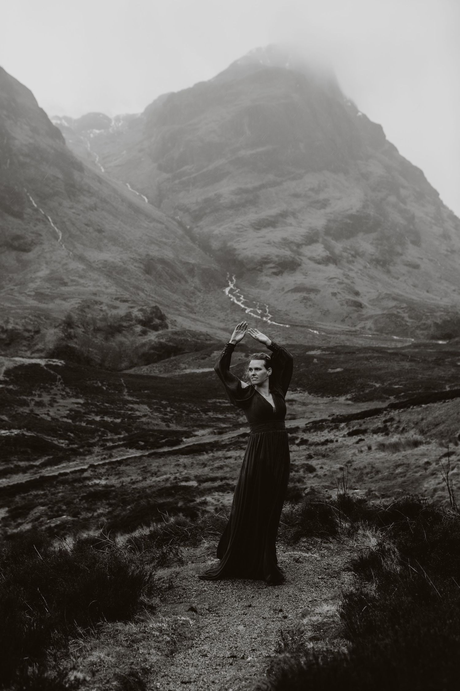 Black and white photo of woman in dark dress holding branches aloft in misty mountain valley.
