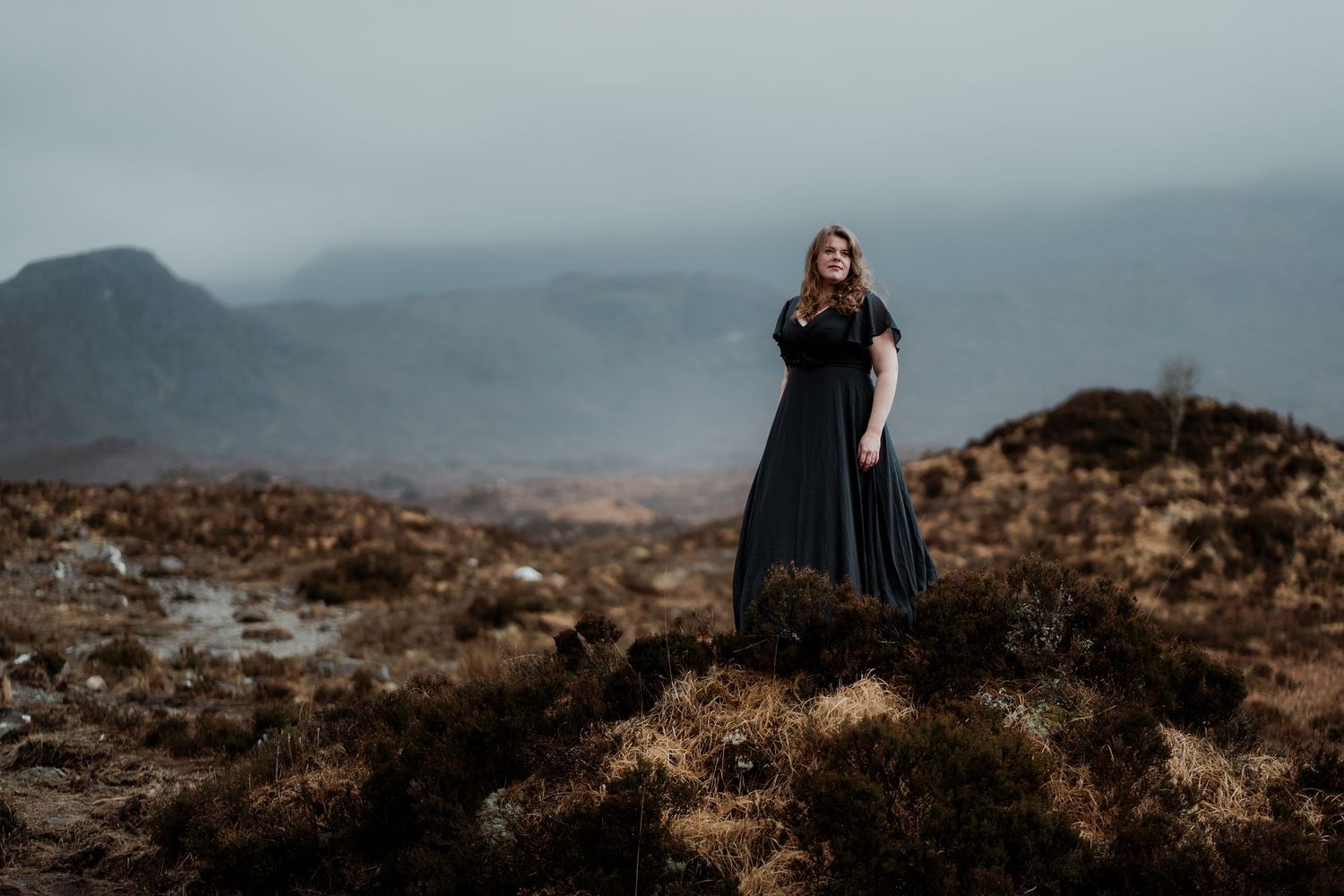 A figure in a flowing dark dress stands on a misty mountainside against a moody atmospheric landscape.