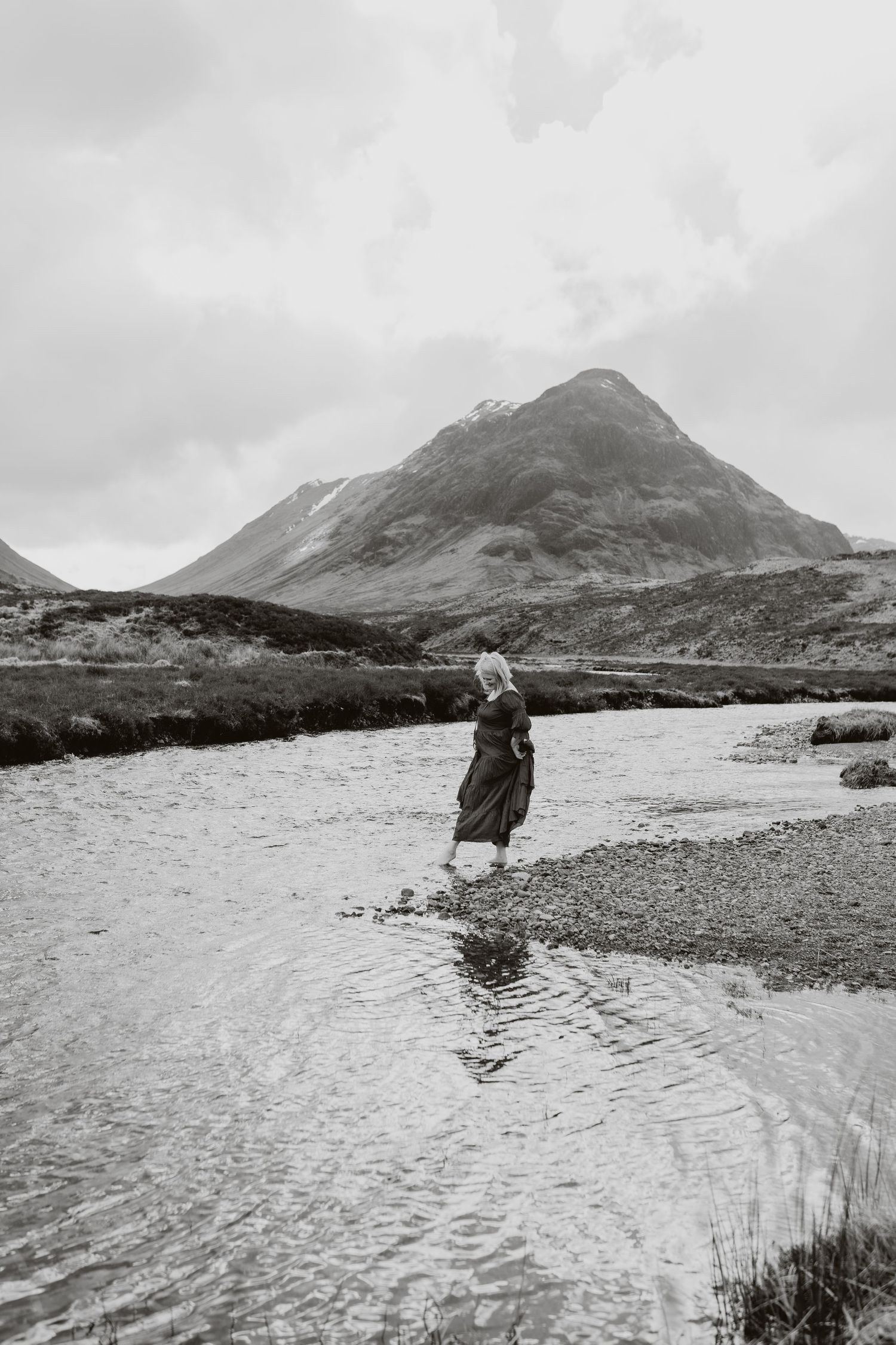 Woman in long dress wading through a shallow river with a dramatic mountain backdrop in black and white.