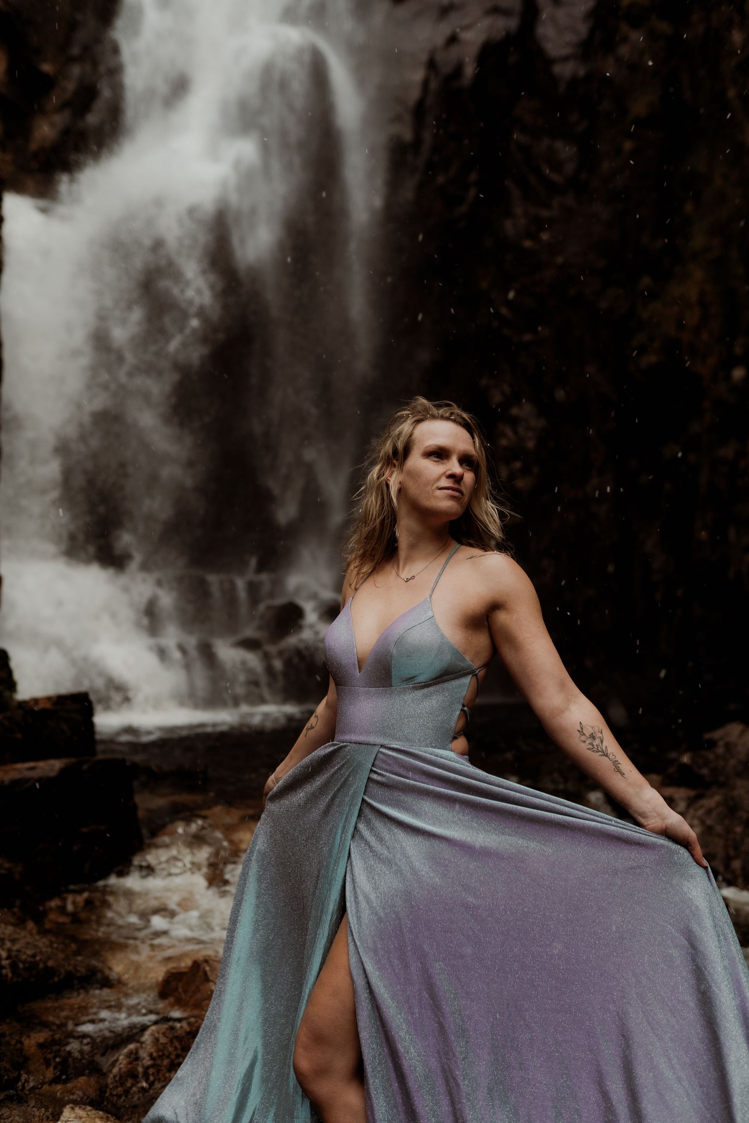 Woman in silver gown posing gracefully in front of a dramatic waterfall surrounded by rocky terrain.