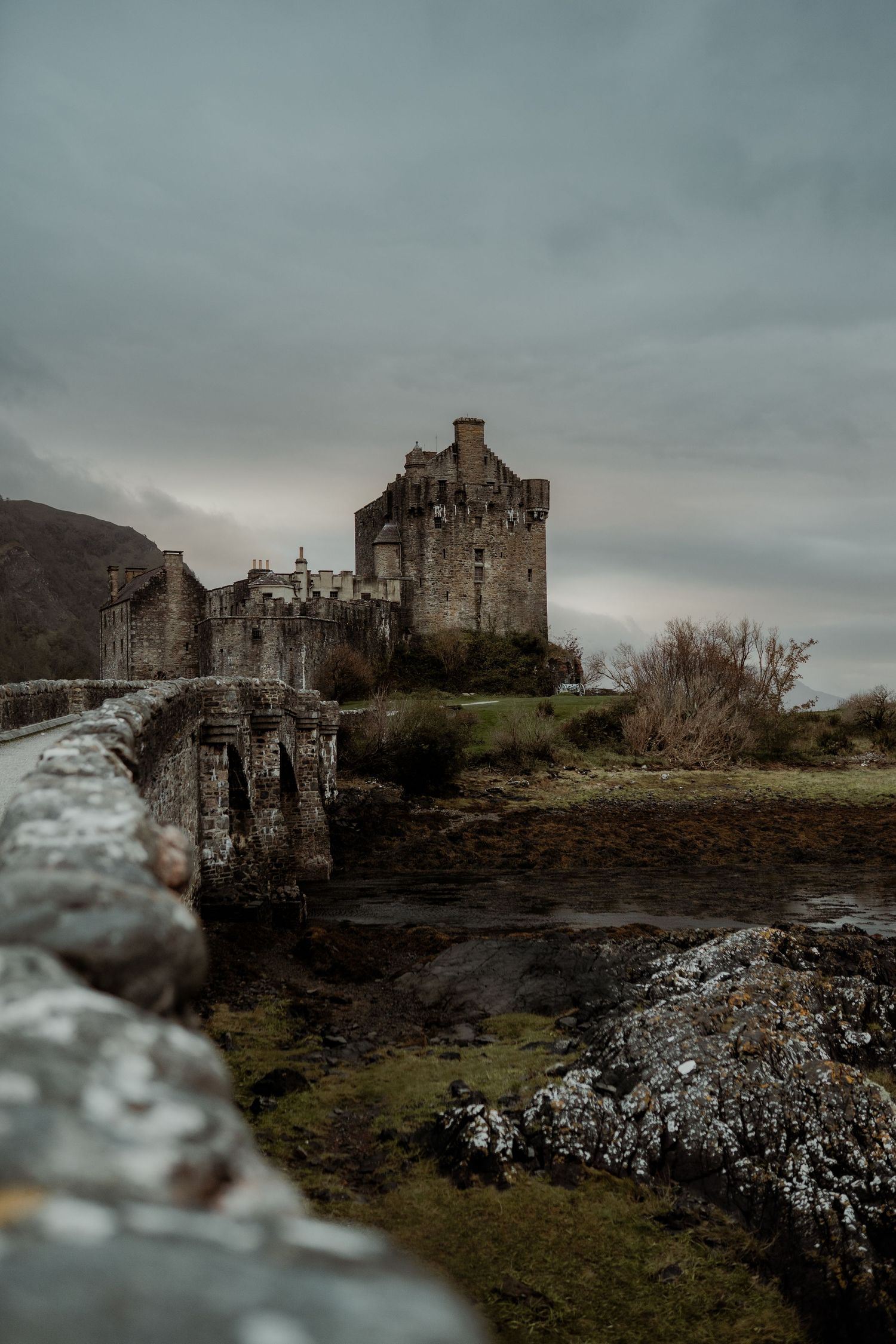 Moody view of Eilean Donan Castle in Scotland on an overcast day with dramatic stormy skies and rocky terrain.