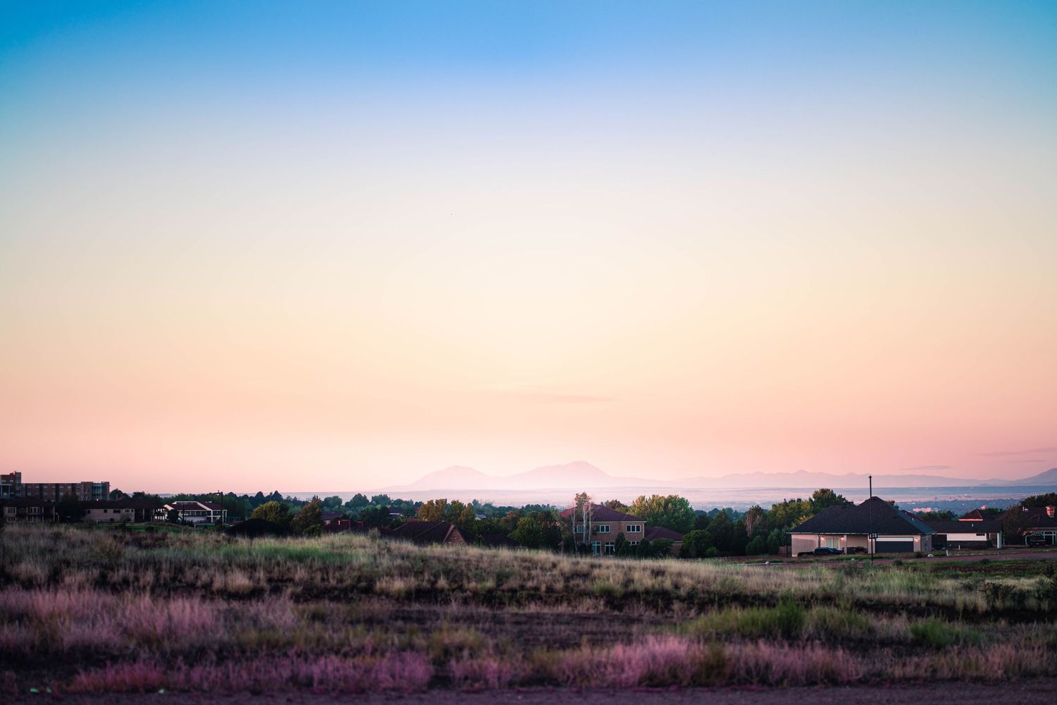 Serene twilight landscape with pink and blue sky over a rural field with scattered trees and buildings at dusk.