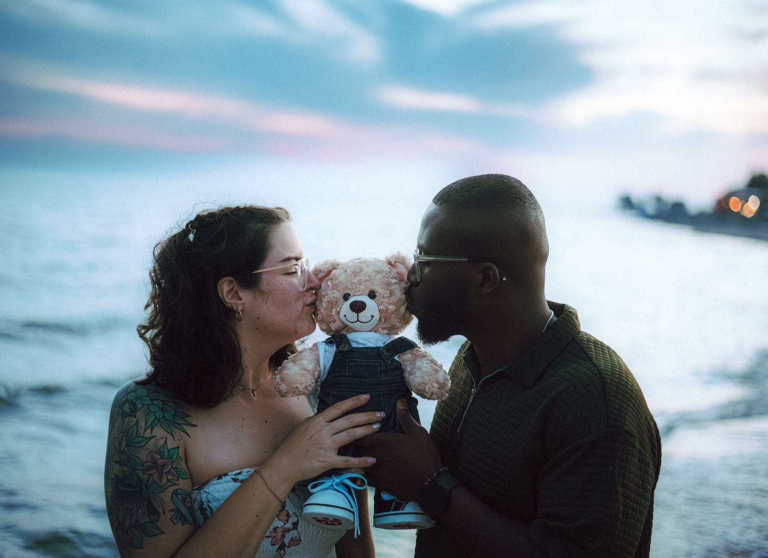 Interracial couple kissing a teddy bear together on a beach at dusk, with ocean waves in the background.