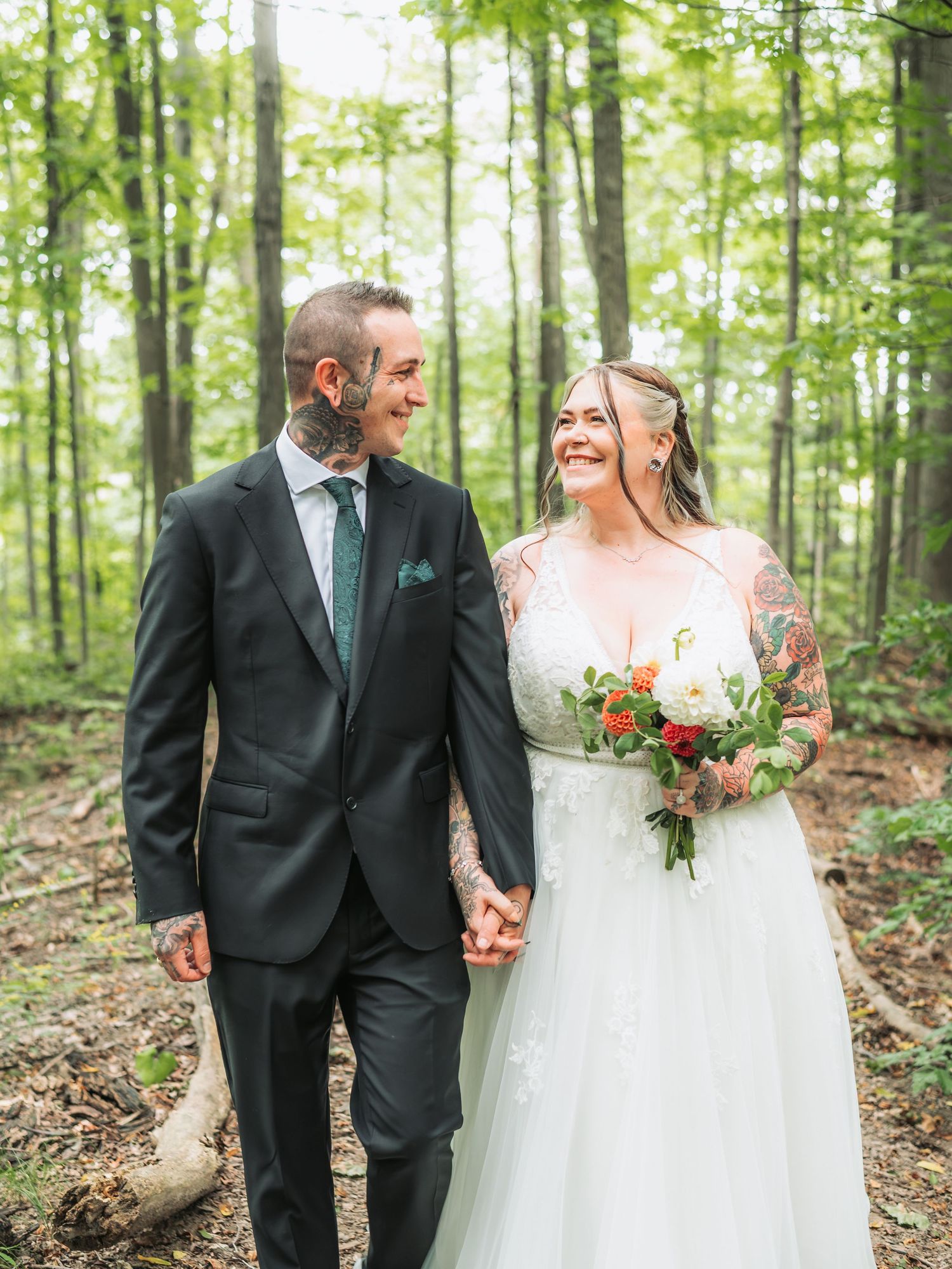 Bride and groom hold hands walking through a forest, she holds a red bouquet, both smiling, he wears a dark suit.
