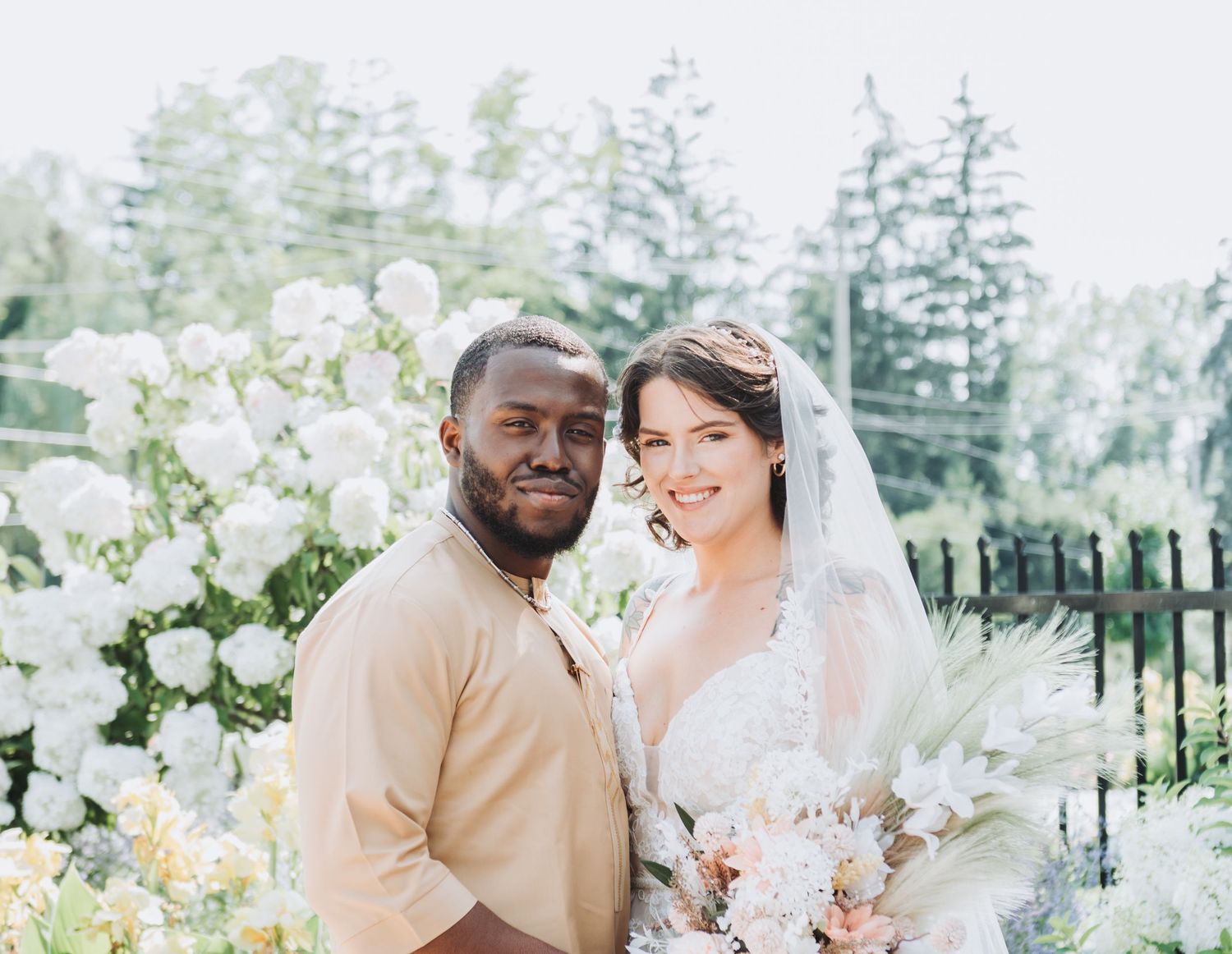 Interracial couple posing together at outdoor wedding ceremony with white floral arrangements and lush greenery backdrop.