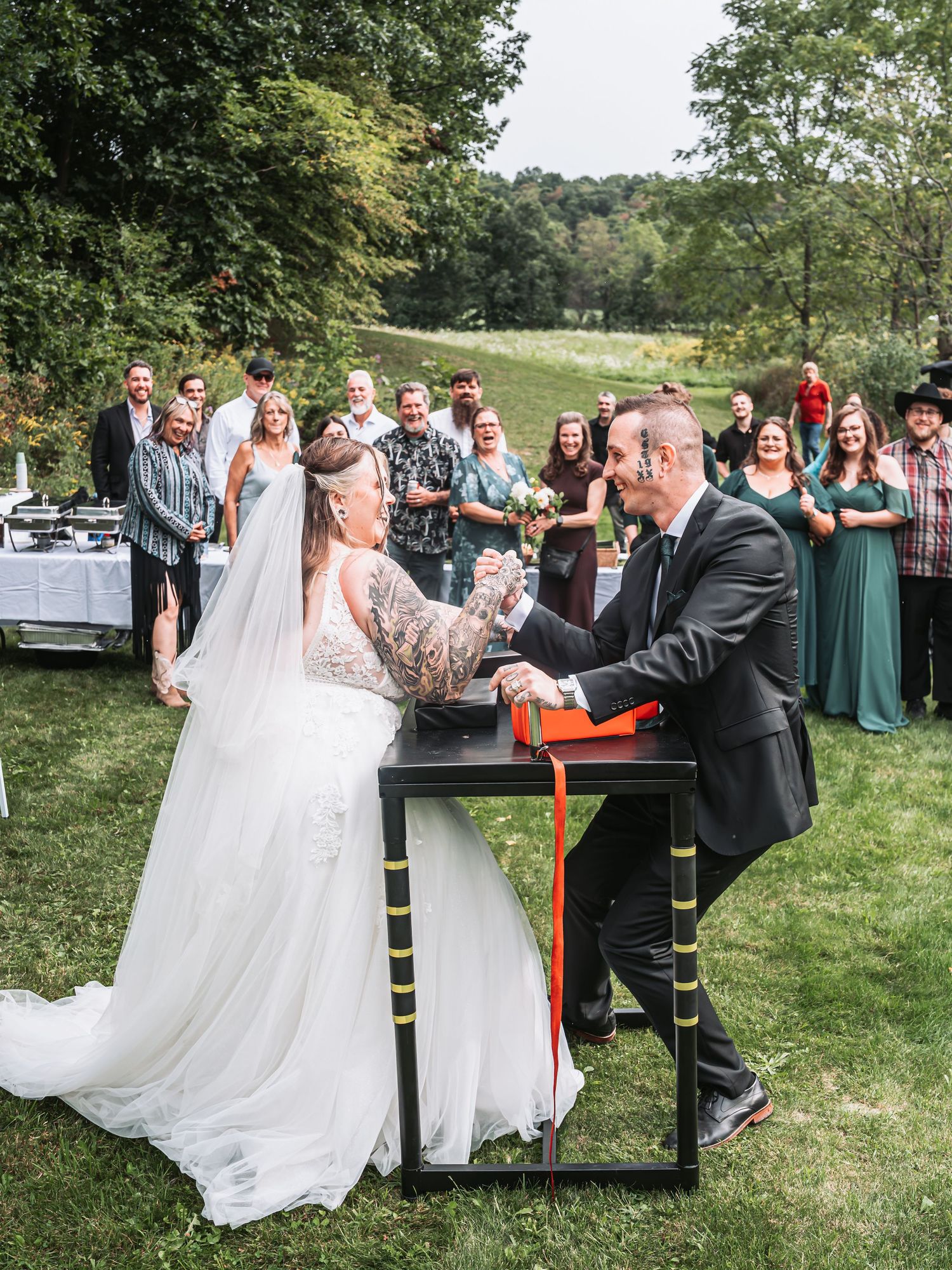 Outdoor wedding ceremony with bride and groom at a decorated table, guests watching on a green hillside.