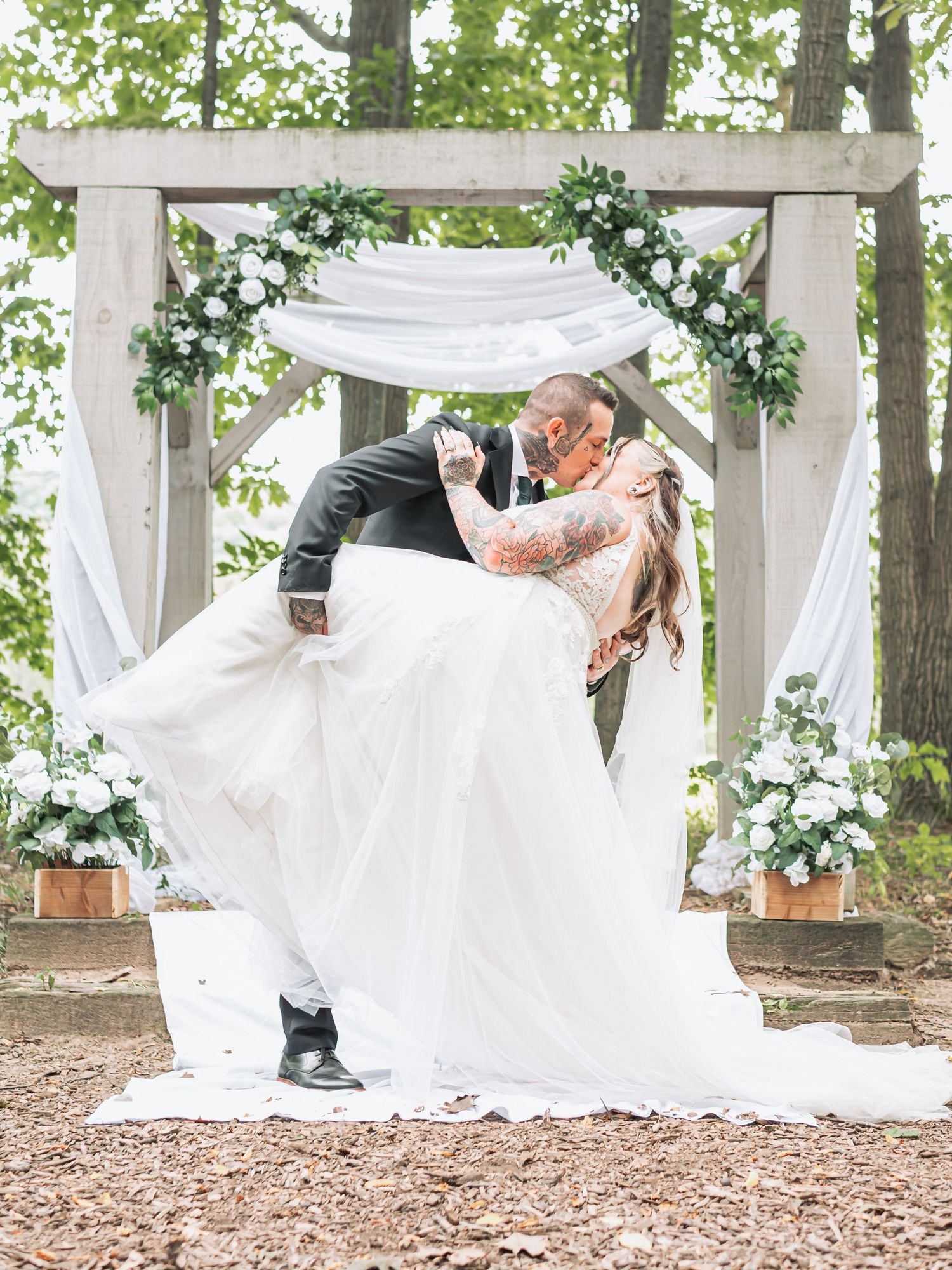 Groom dips bride for a kiss under a rustic wooden arch adorned with white flowers and greenery at an outdoor wedding.